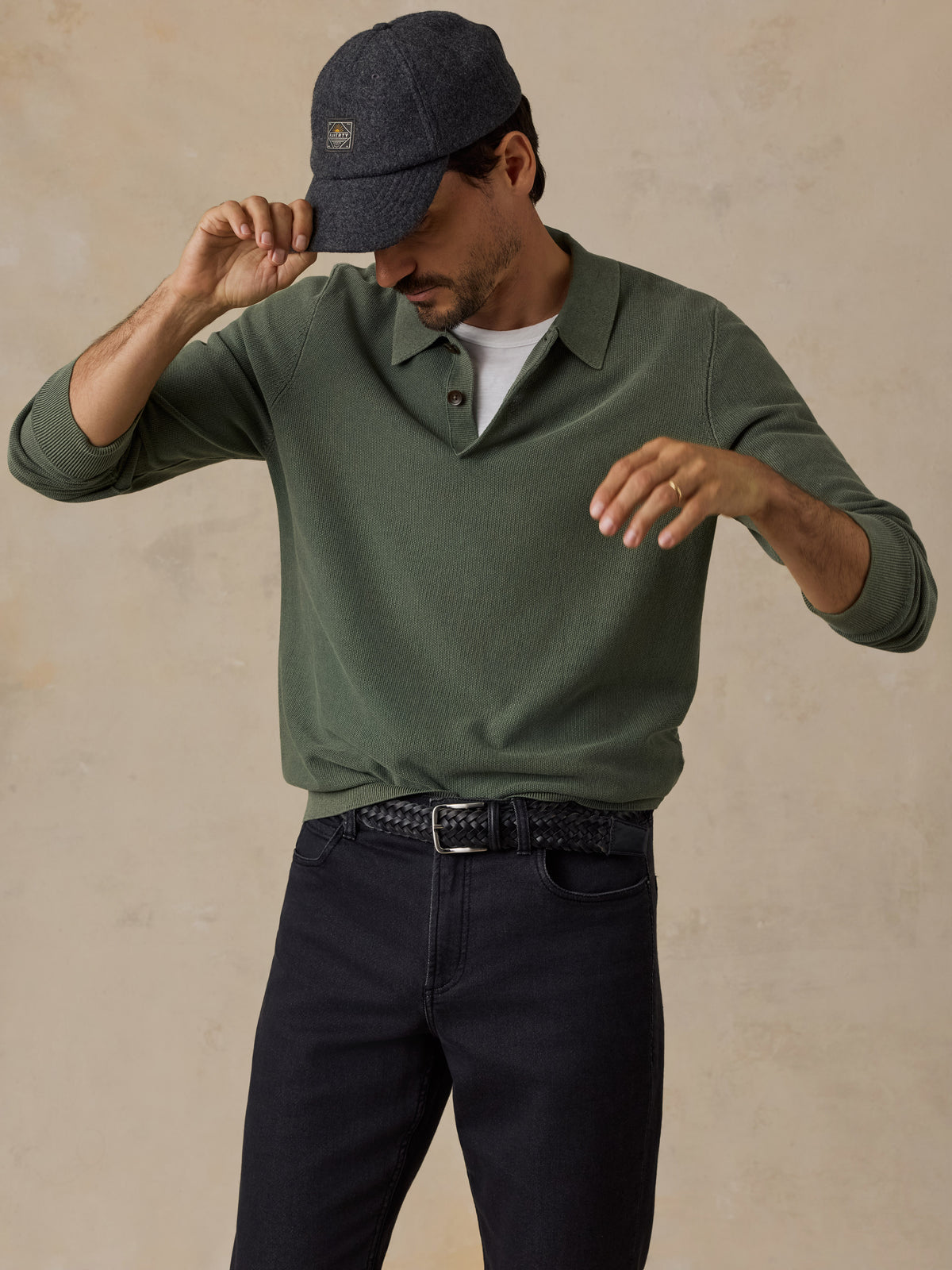 A man wearing an olive green long-sleeve polo shirt, dark navy pants, a dark grey cap, and a white undershirt, posing against a textured beige background., image 5 of 7