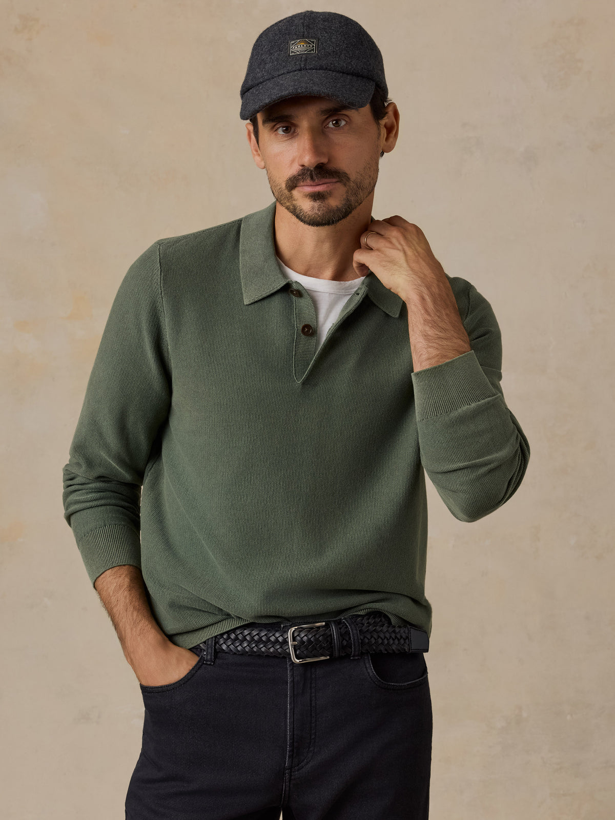 A man wearing an olive green long-sleeve polo shirt, a dark grey baseball cap, a white undershirt, and dark jeans, standing against a textured beige background., image 1 of 7