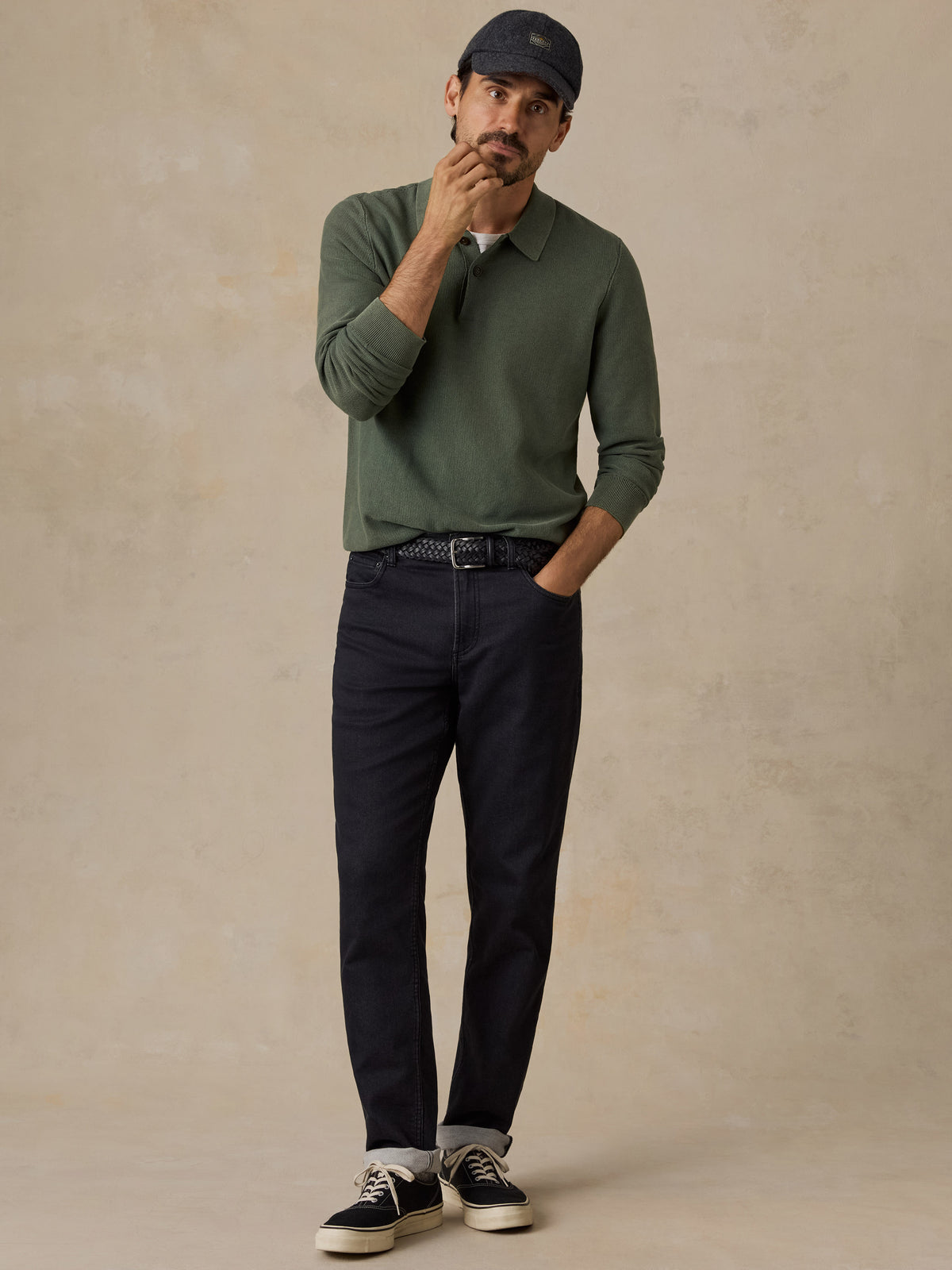 A man wearing an olive green long-sleeve polo shirt, dark navy pants, a navy baseball cap, and black sneakers with white soles, standing against a textured beige studio backdrop., image 7 of 7