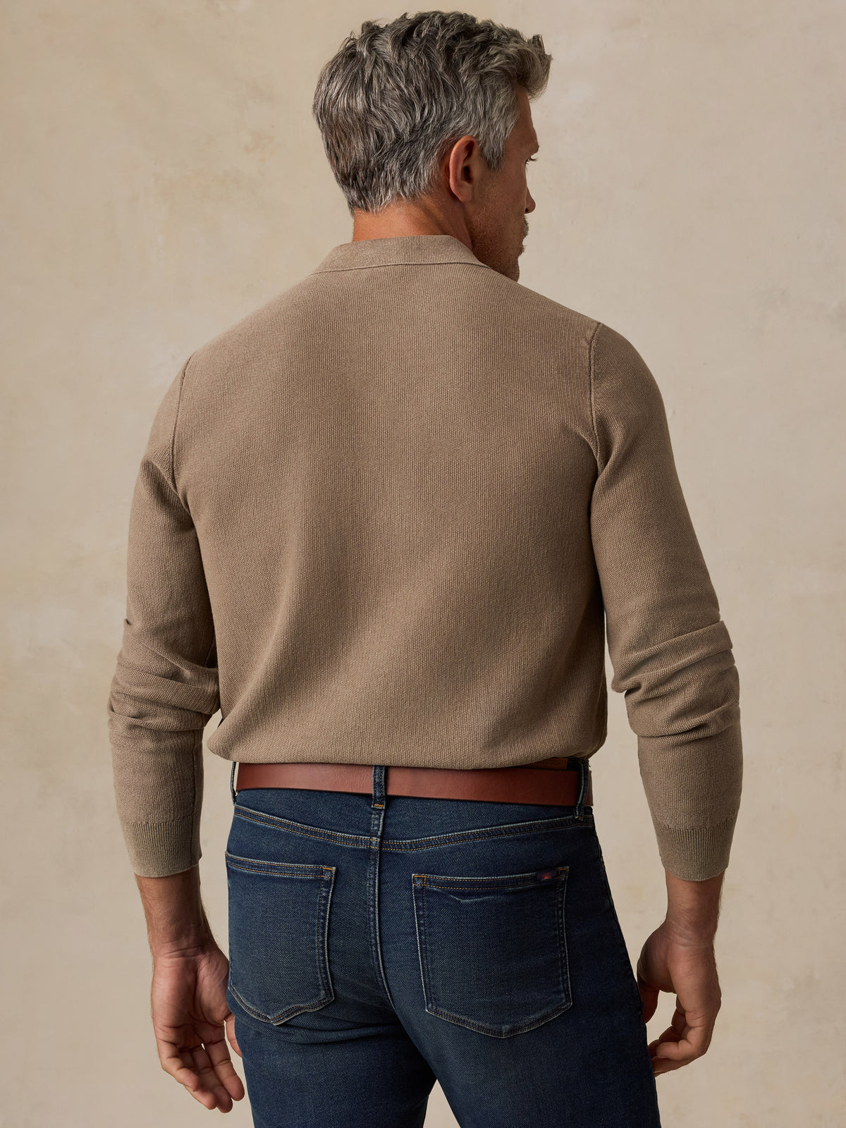 A man viewed from behind wearing a taupe brown knit sweater and dark blue jeans, standing against a textured beige background., image 3 of 5