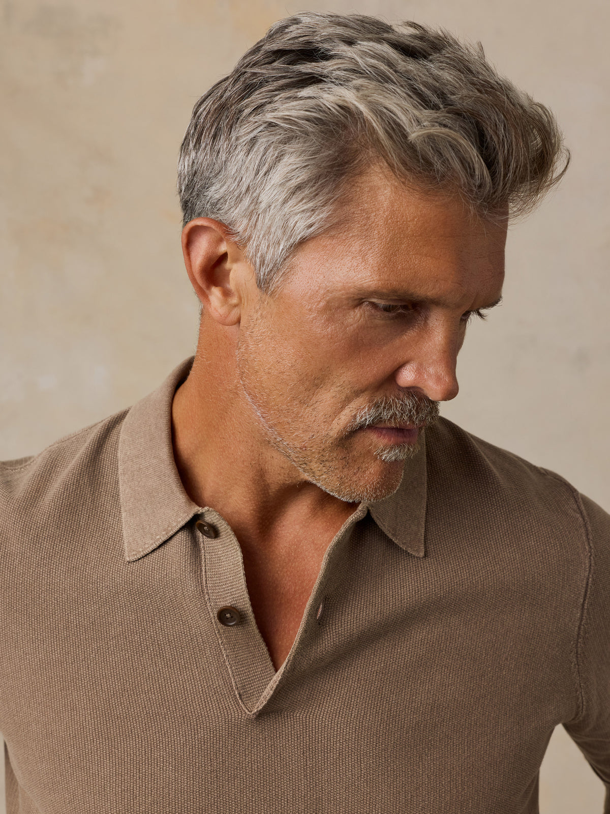 A close-up of a man wearing a brown ribbed knit polo shirt, looking down and to the side against a textured beige background., image 4 of 5