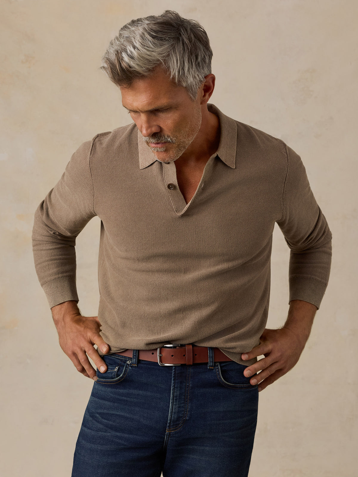 A man with gray hair wearing a taupe brown long-sleeve knit polo shirt and dark blue jeans, standing against a textured beige background., image 1 of 5