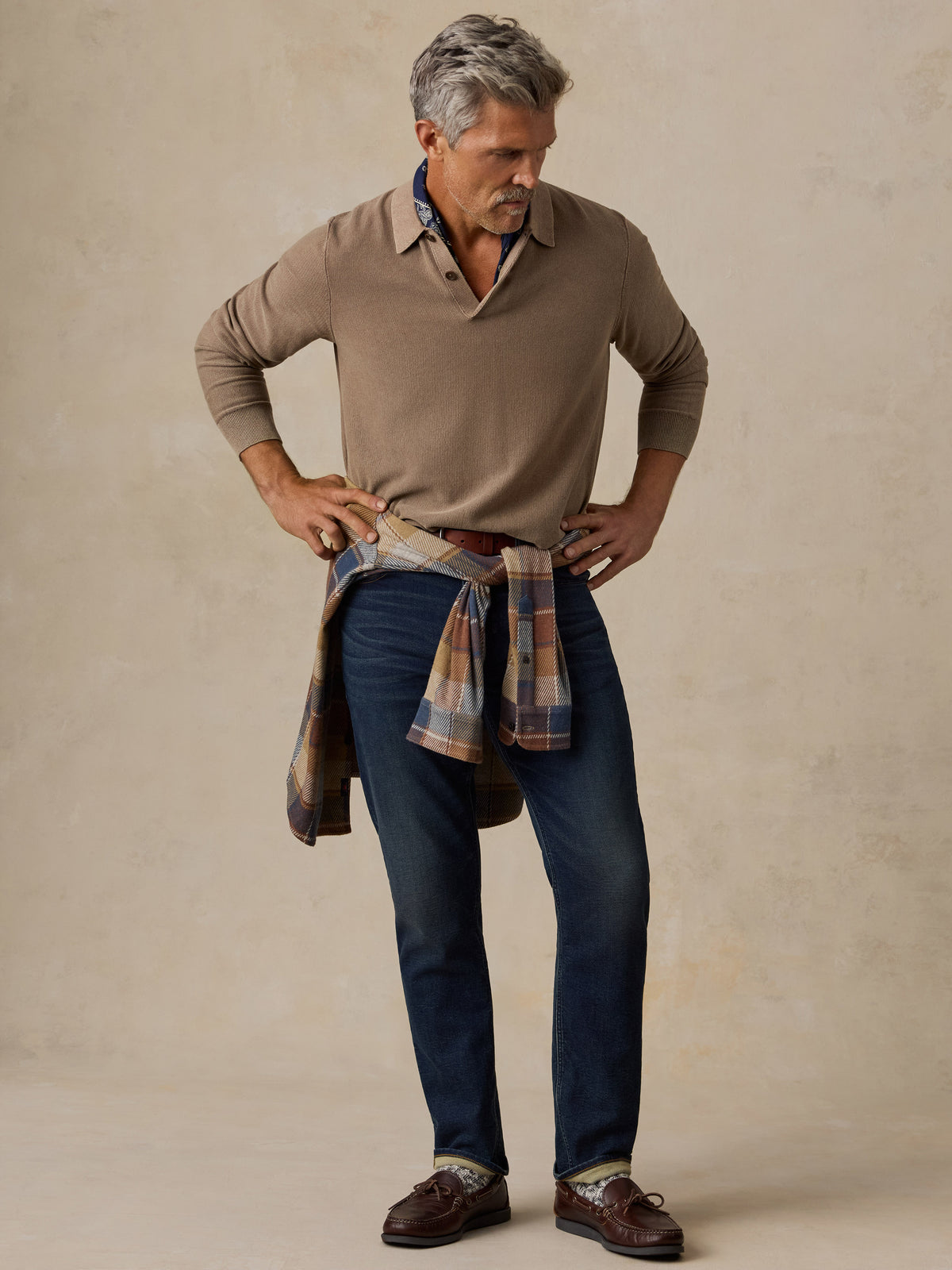 A man modeling a brown long-sleeve knit polo shirt, dark blue jeans, and brown leather shoes, with a plaid shirt tied around his waist, against a textured beige studio backdrop., image 2 of 5