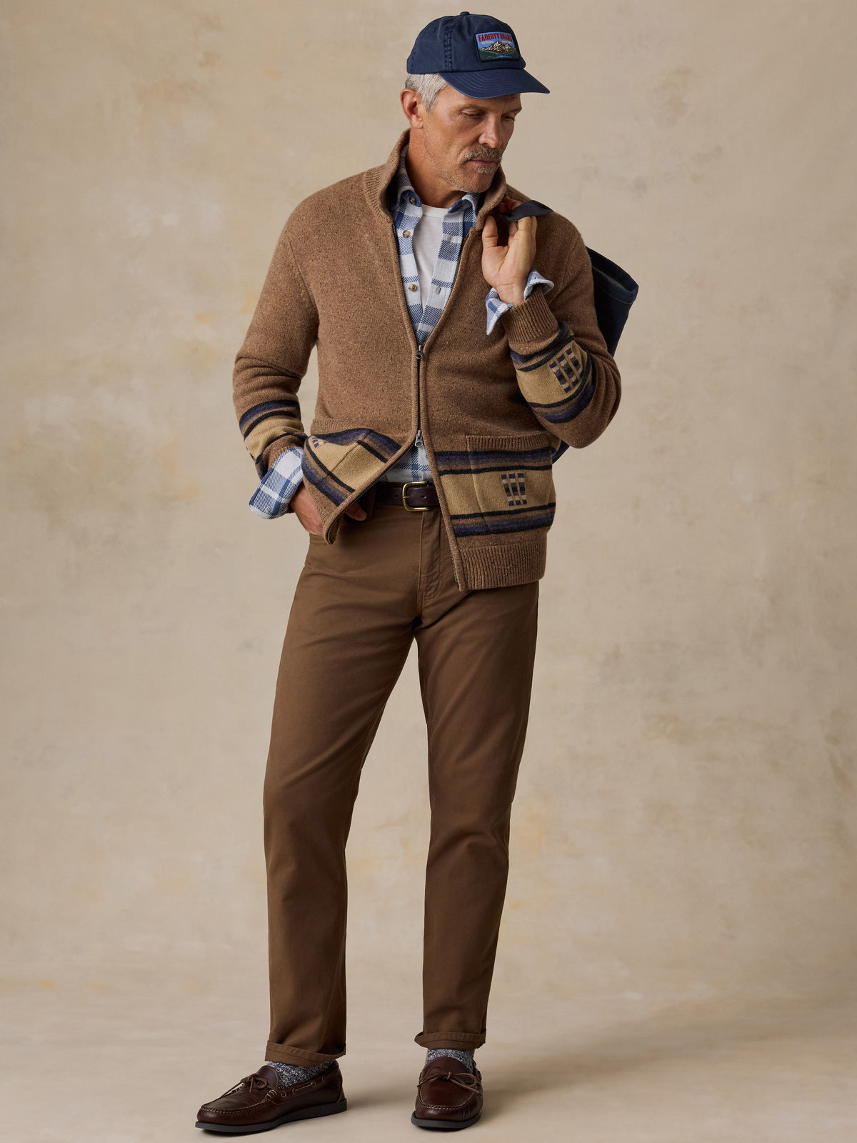 A man modeling a brown knit zip-up cardigan with striped cuffs and hem, paired with brown trousers, a blue plaid shirt, a navy baseball cap, and brown leather shoes against a textured beige studio backdrop., image 5 of 7