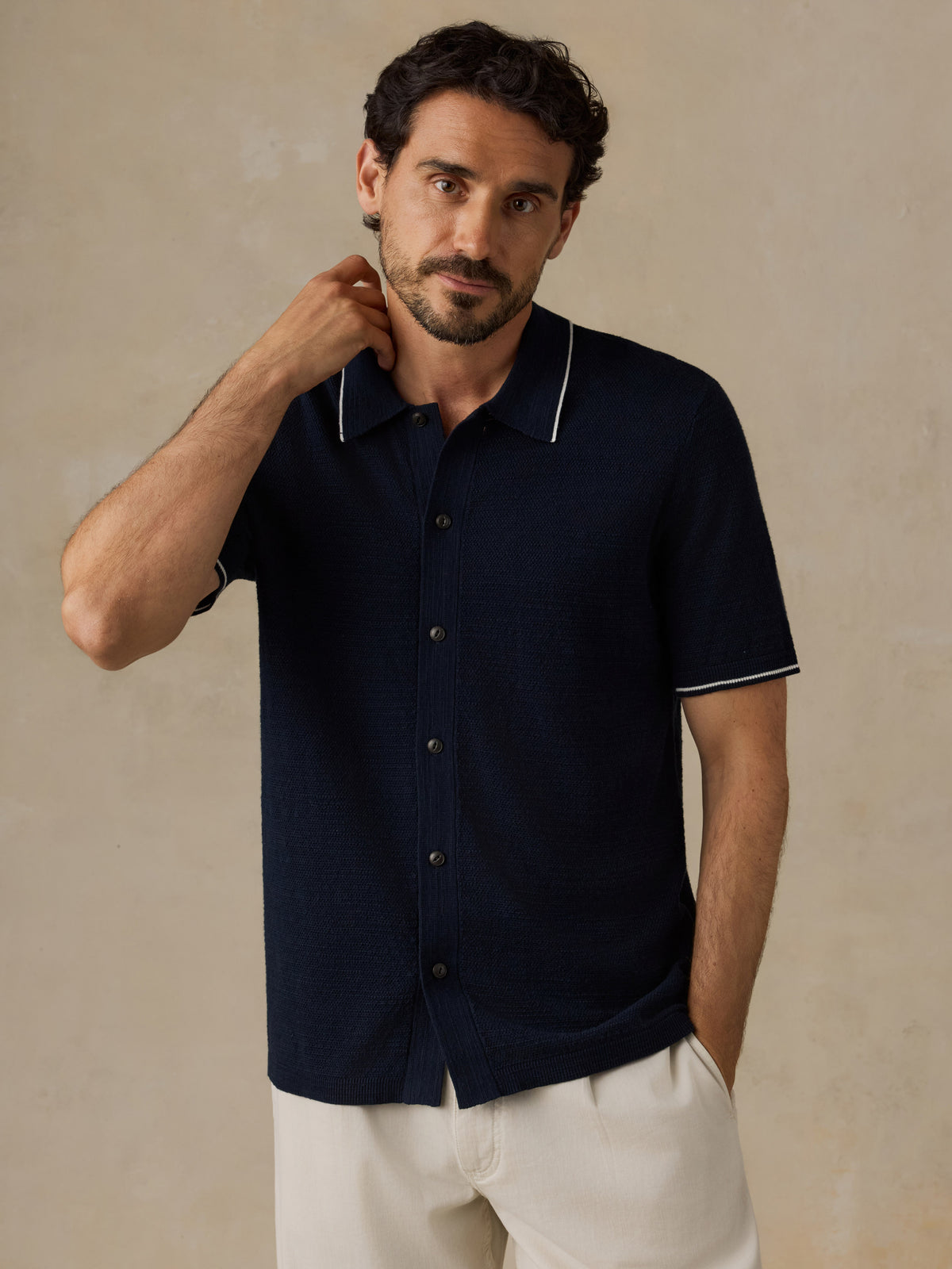 A man wearing a dark navy short-sleeve knit polo shirt with white piping on the collar and sleeve cuffs, paired with off-white trousers, standing against a textured beige background., image 2 of 6