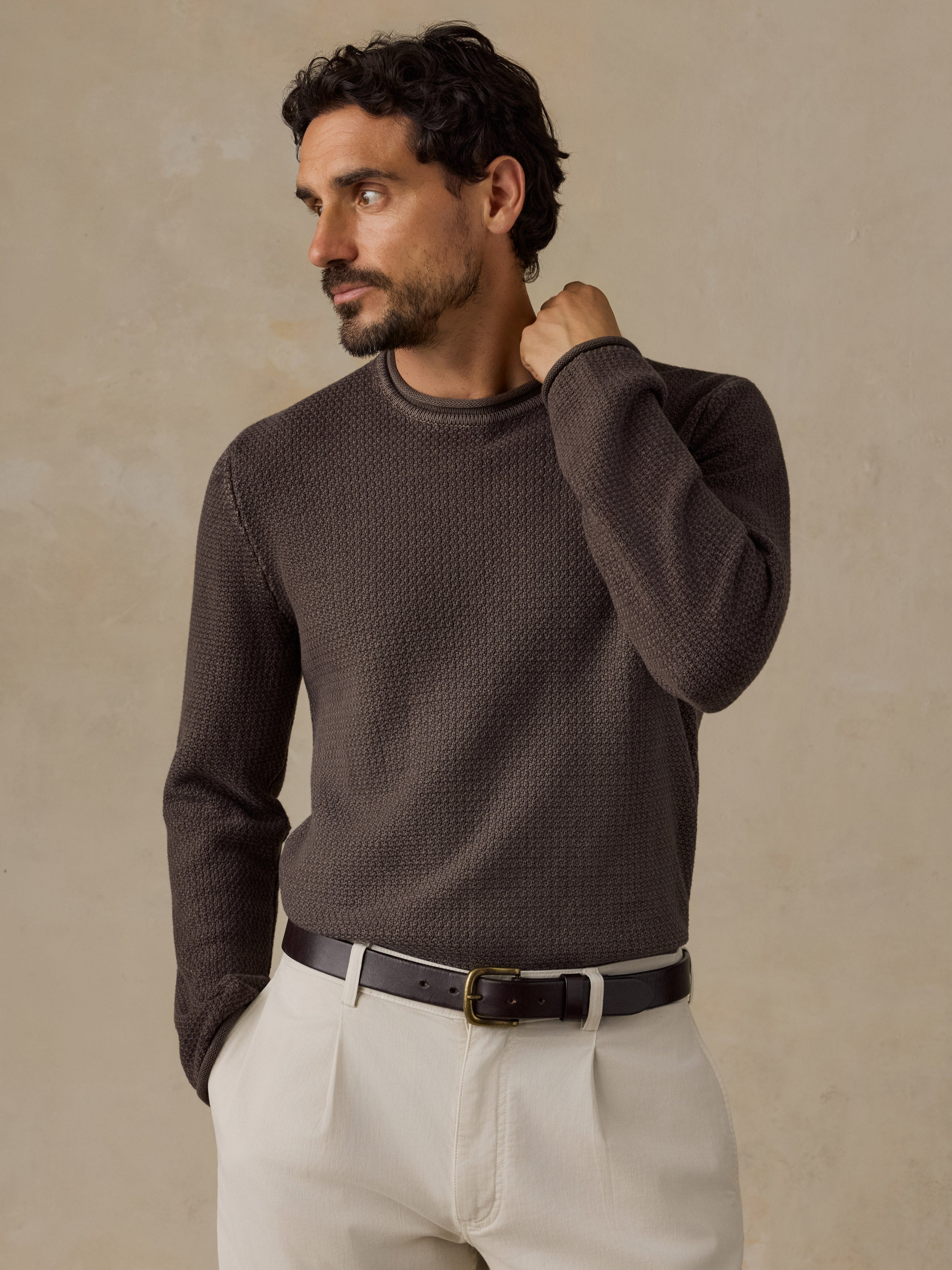 A man wearing a dark brown textured knit crewneck sweater, cream-colored trousers, and a black leather belt, posing against a warm beige textured background.