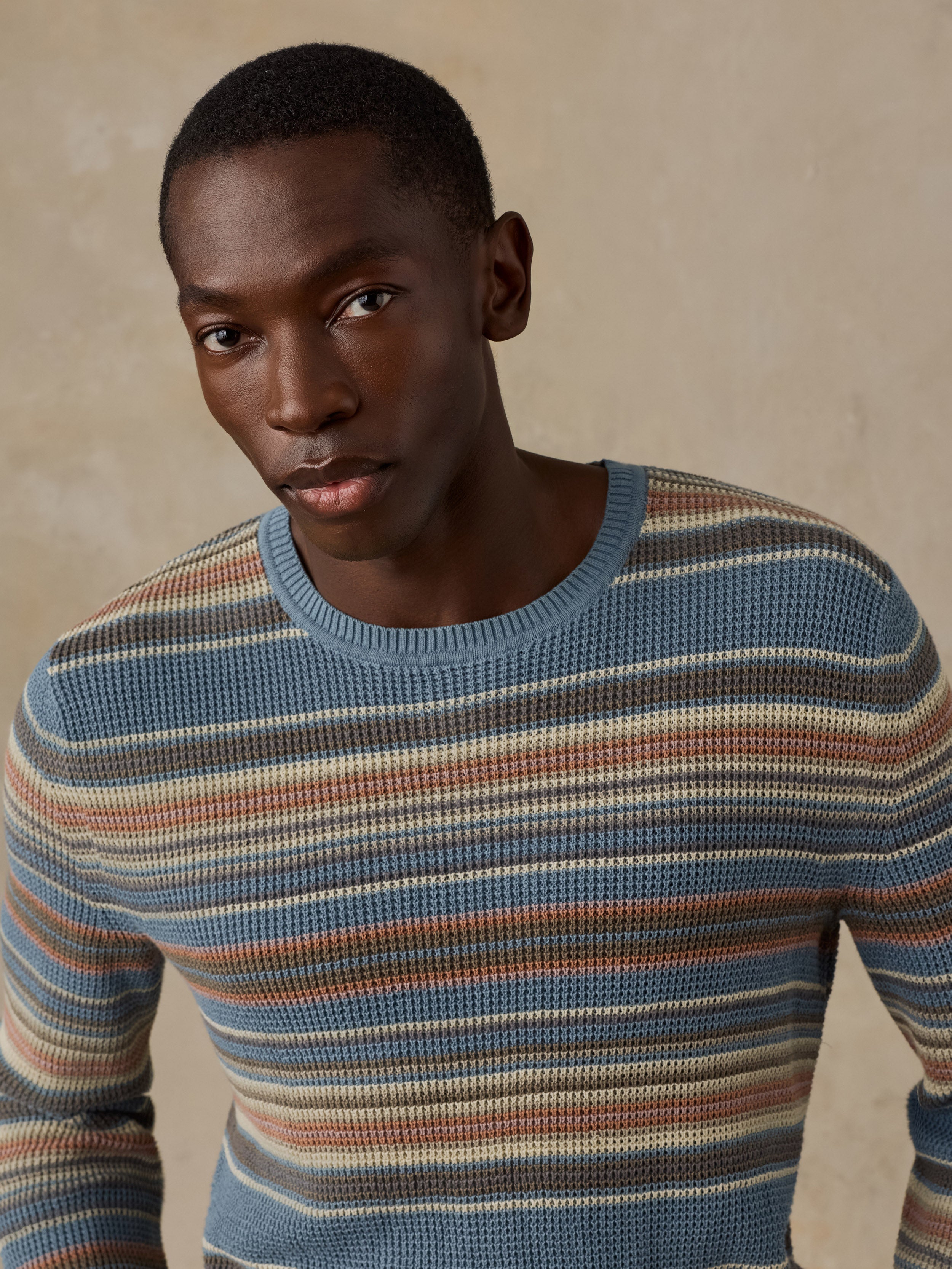 A man wearing a blue, beige, and brown horizontally striped crewneck sweater standing against a beige background.