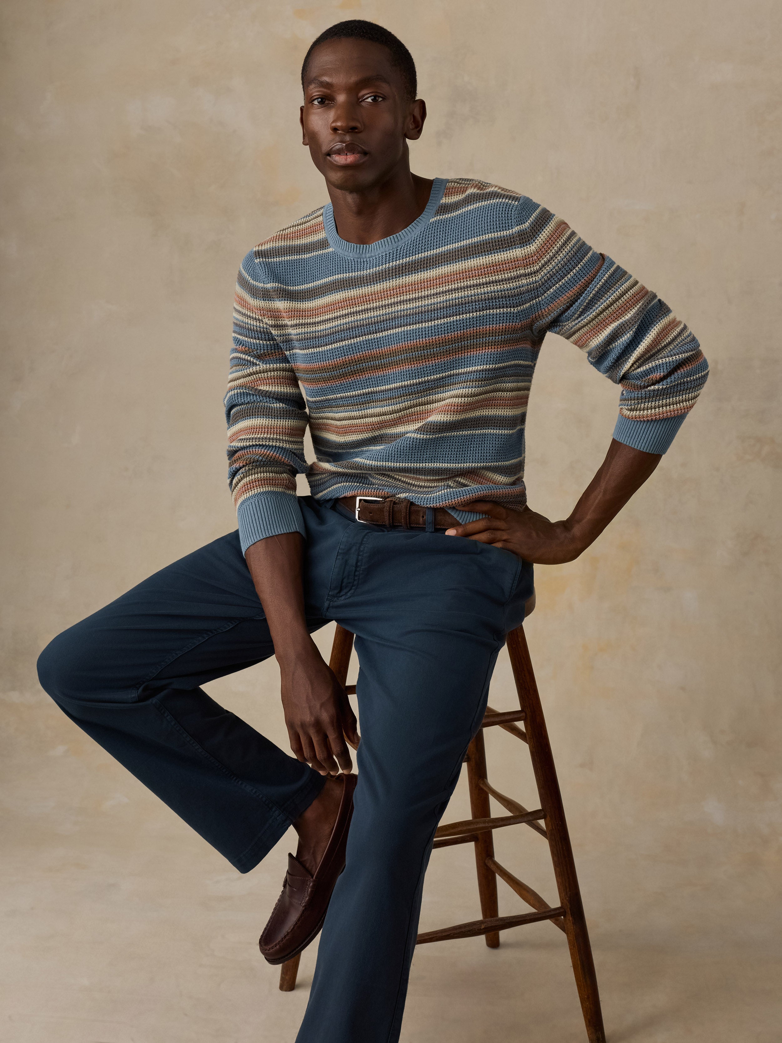 A man sitting on a wooden stool wearing a blue, beige, and brown horizontally striped crewneck sweater and dark navy trousers, standing against a beige background.