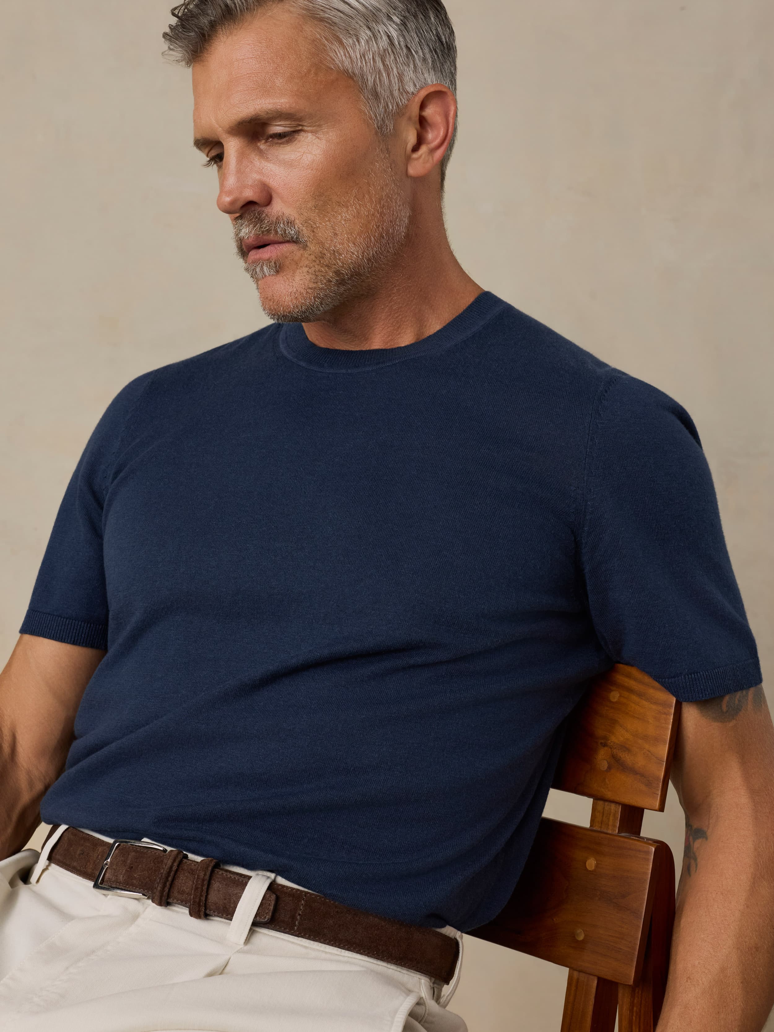 Man wearing a navy blue t-shirt sitting on a wooden chair against a beige background
