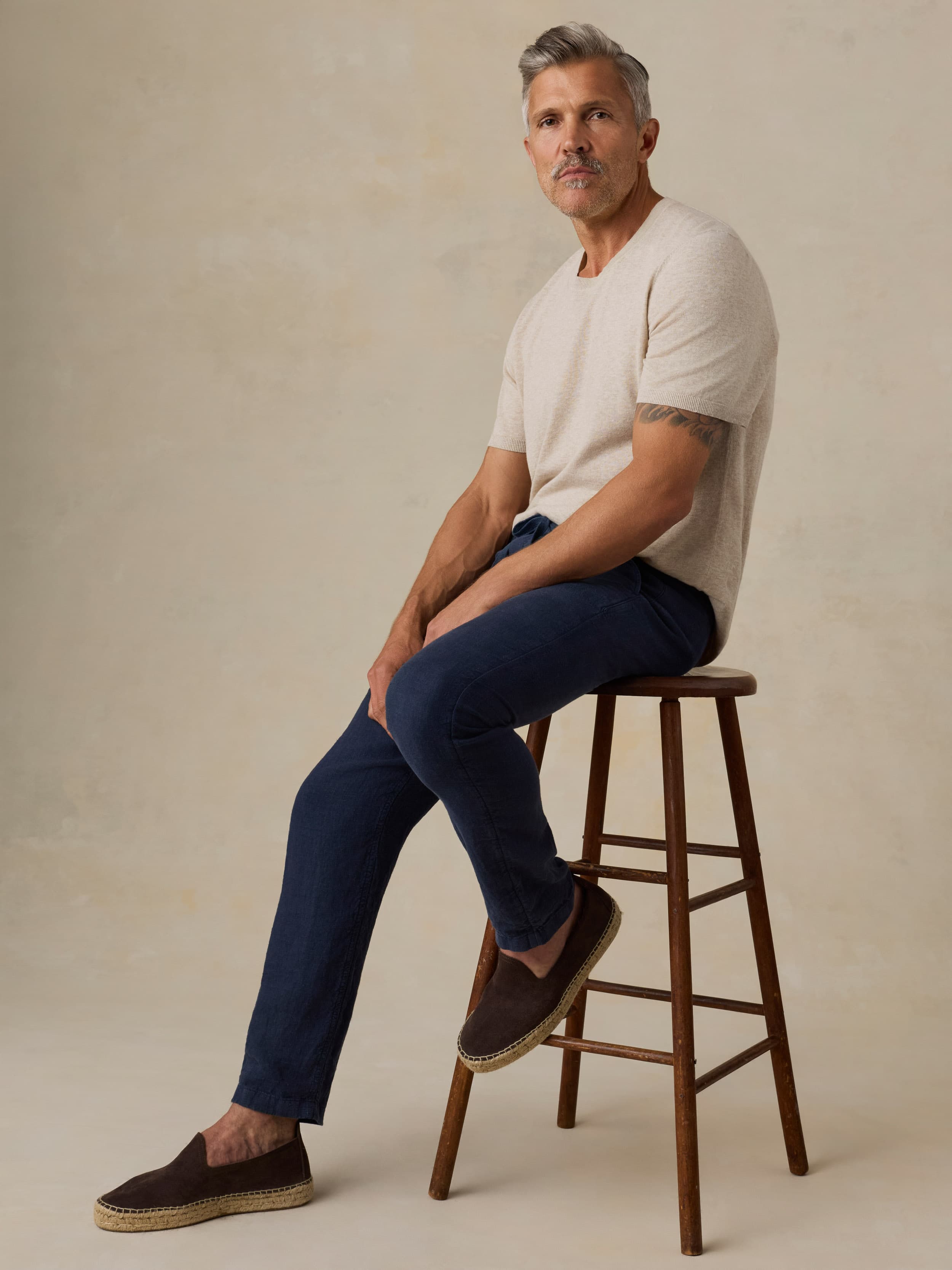 Man sitting on a wooden stool wearing a beige t-shirt and navy blue pants against a beige background
