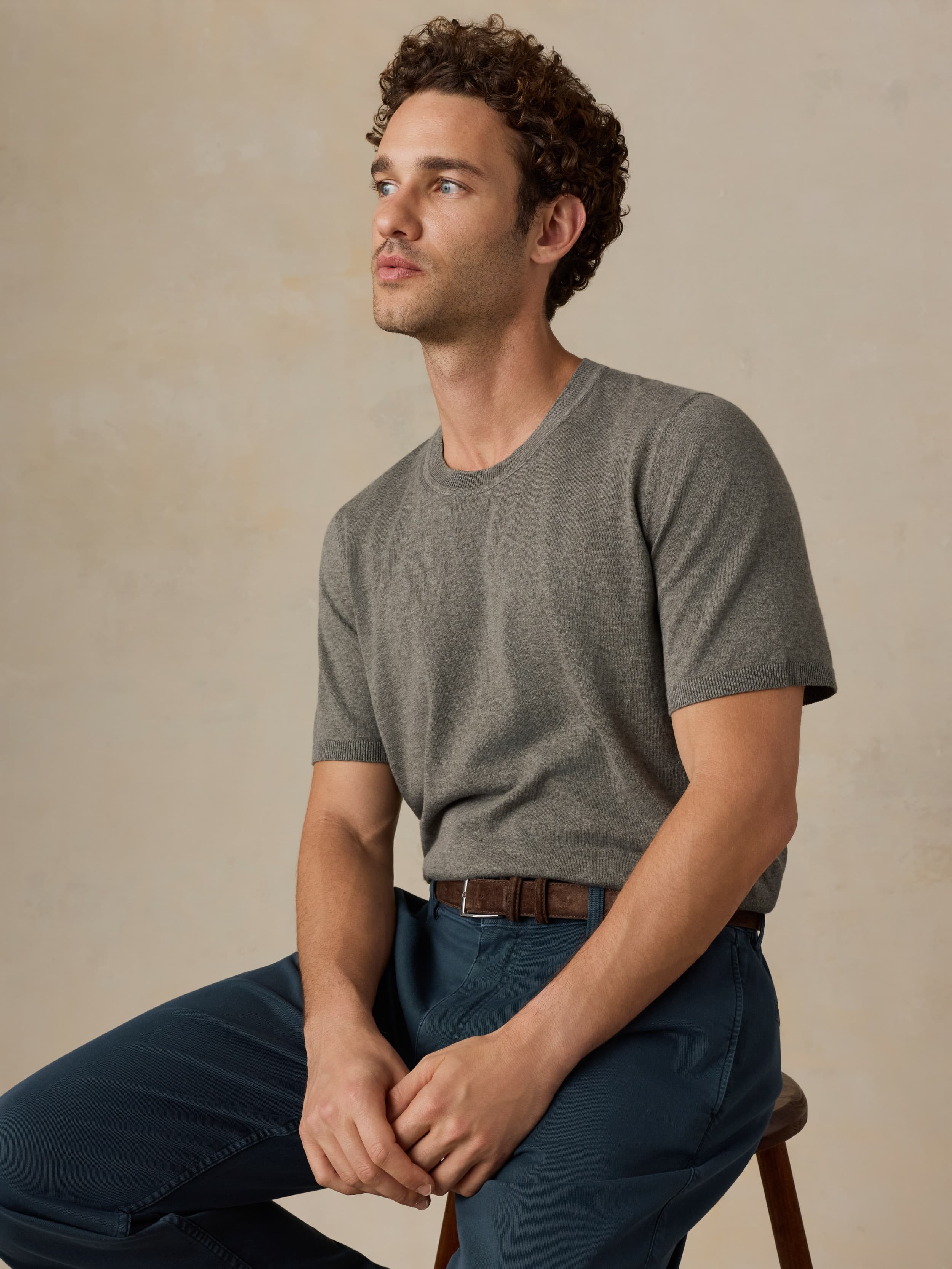 Man wearing a green t-shirt and navy blue pants sitting on a wooden stool against a beige background