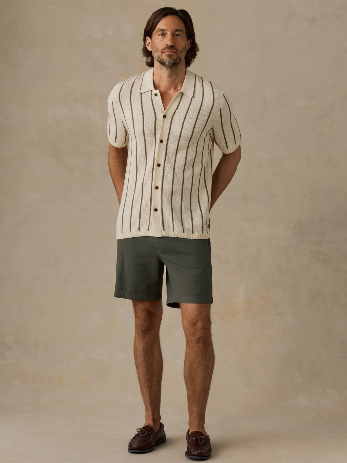 A man modeling a short-sleeve button-up shirt with thin vertical black stripes on an off-white base, paired with dark olive green shorts and brown leather shoes, against a textured beige studio backdrop., image 5 of 6