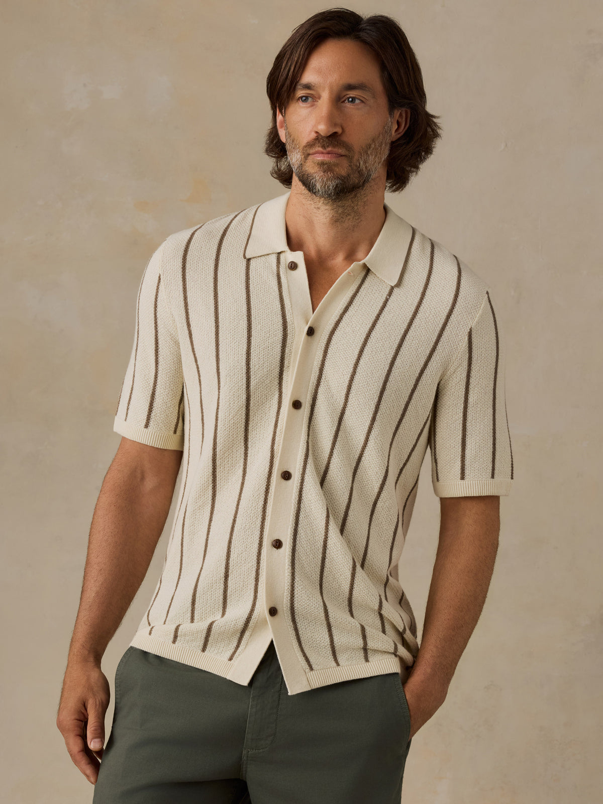 A man wearing a cream-colored short-sleeve knit button-up shirt with vertical brown stripes, paired with brown trousers, standing against a neutral beige background., image 1 of 6
