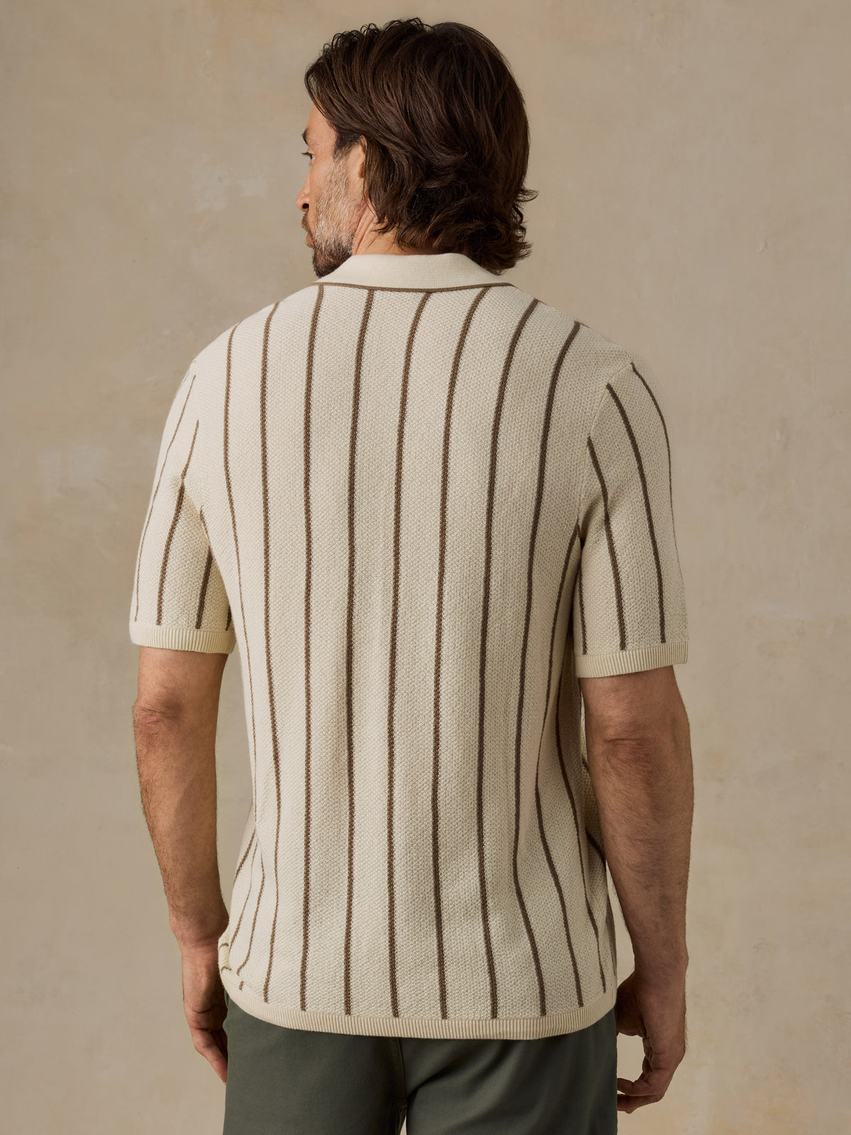 A man viewed from the back wearing a cream knit short-sleeve button-up shirt with vertical brown stripes, paired with brown trousers, against a textured beige background., image 3 of 6