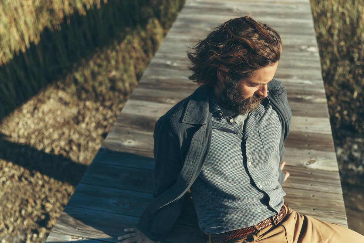 Male model sitting on a dock at sunset wearing an indigo chore coat, a coastline knit shirt and twill pants