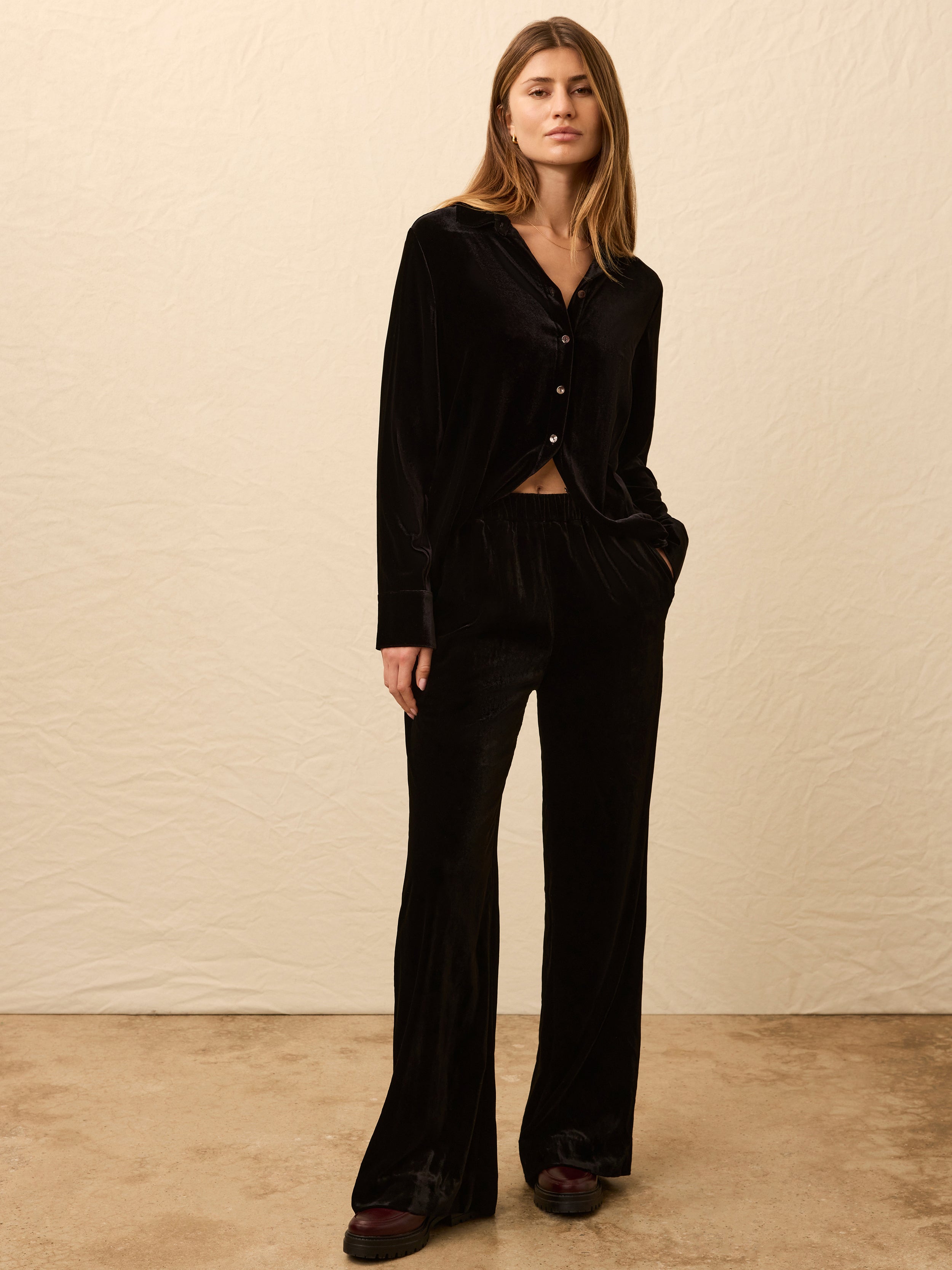 Woman is wearing a black velvet long sleeve button down shirt with matching pants against a beige background