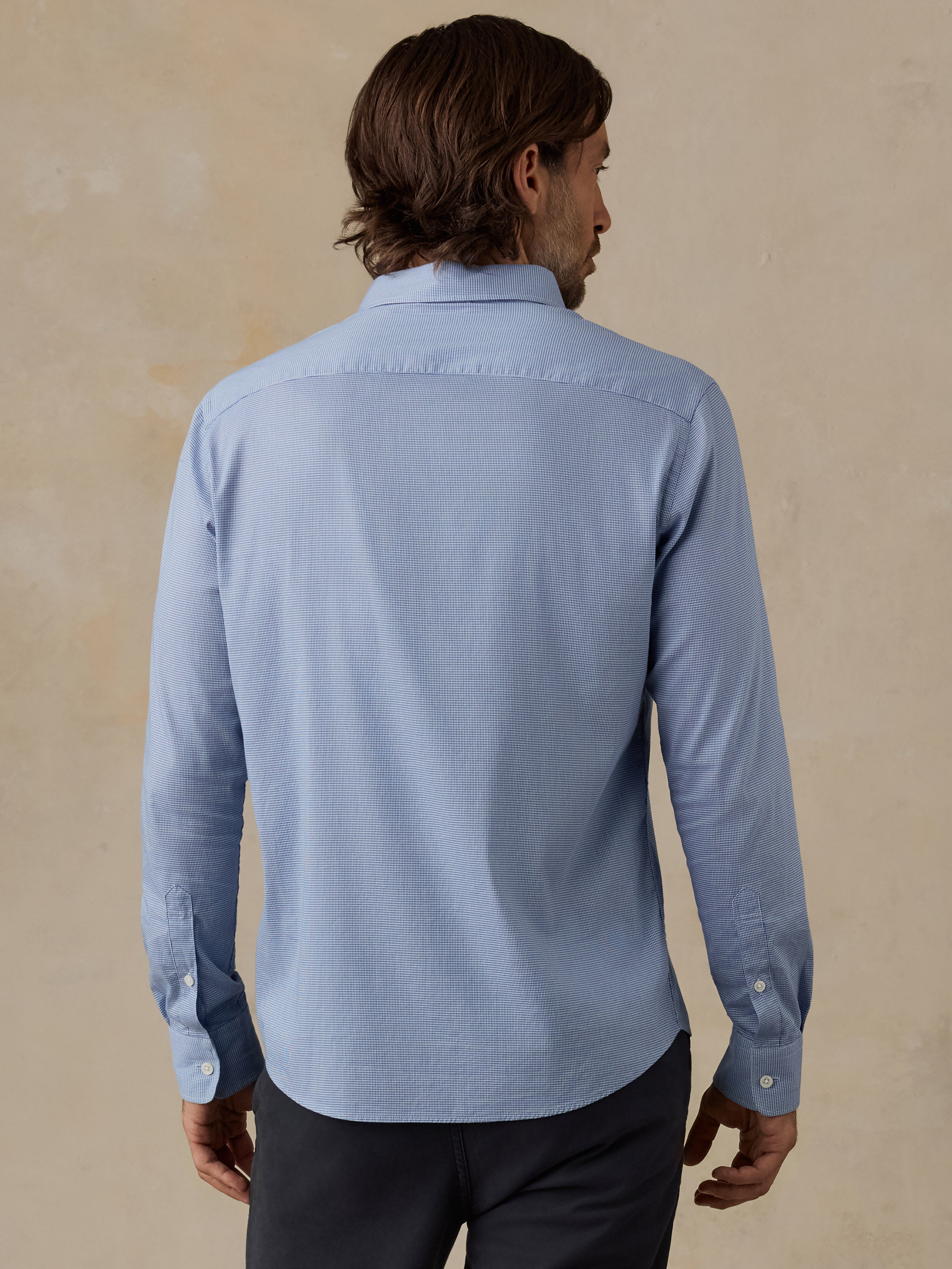 A man viewed from the back wearing a light blue long-sleeve button-up shirt and dark trousers, standing against a textured beige wall.