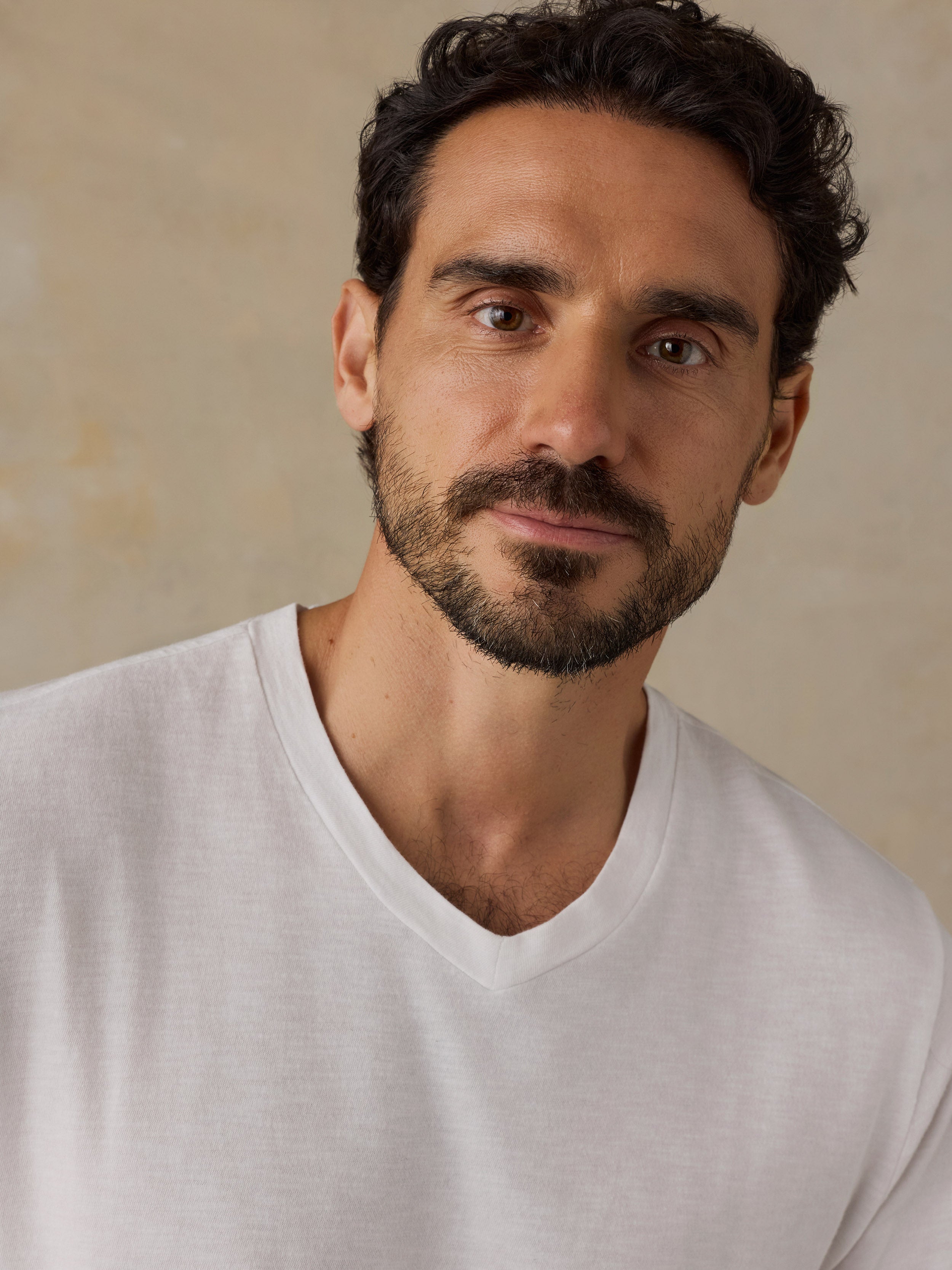 A close-up portrait of a man with dark curly hair and a beard, wearing a white v-neck t-shirt against a neutral beige background.