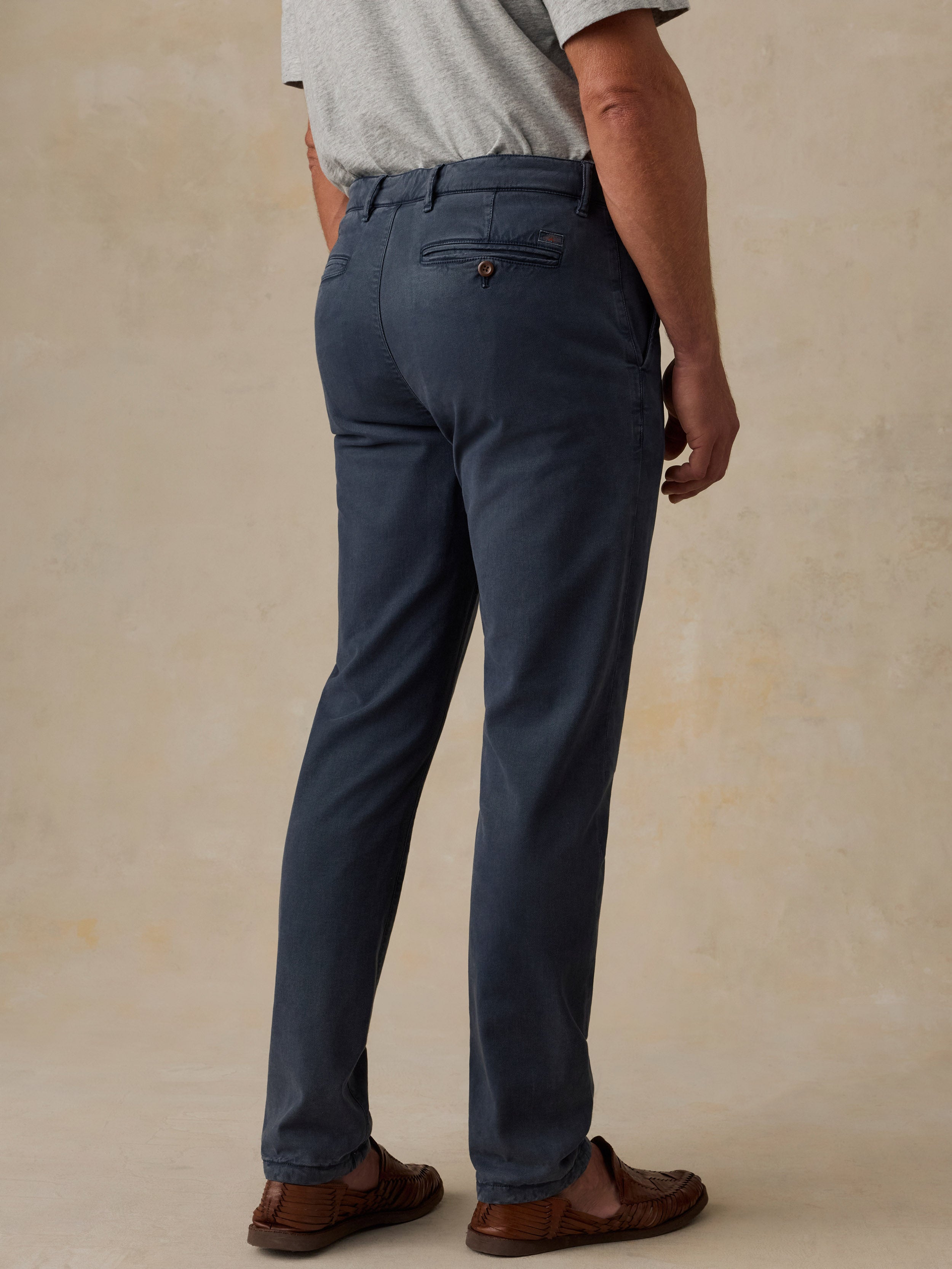 A man wearing navy blue chino pants, a light gray t-shirt, and brown leather loafers, shown from a rear three-quarter angle against a textured beige studio backdrop.
