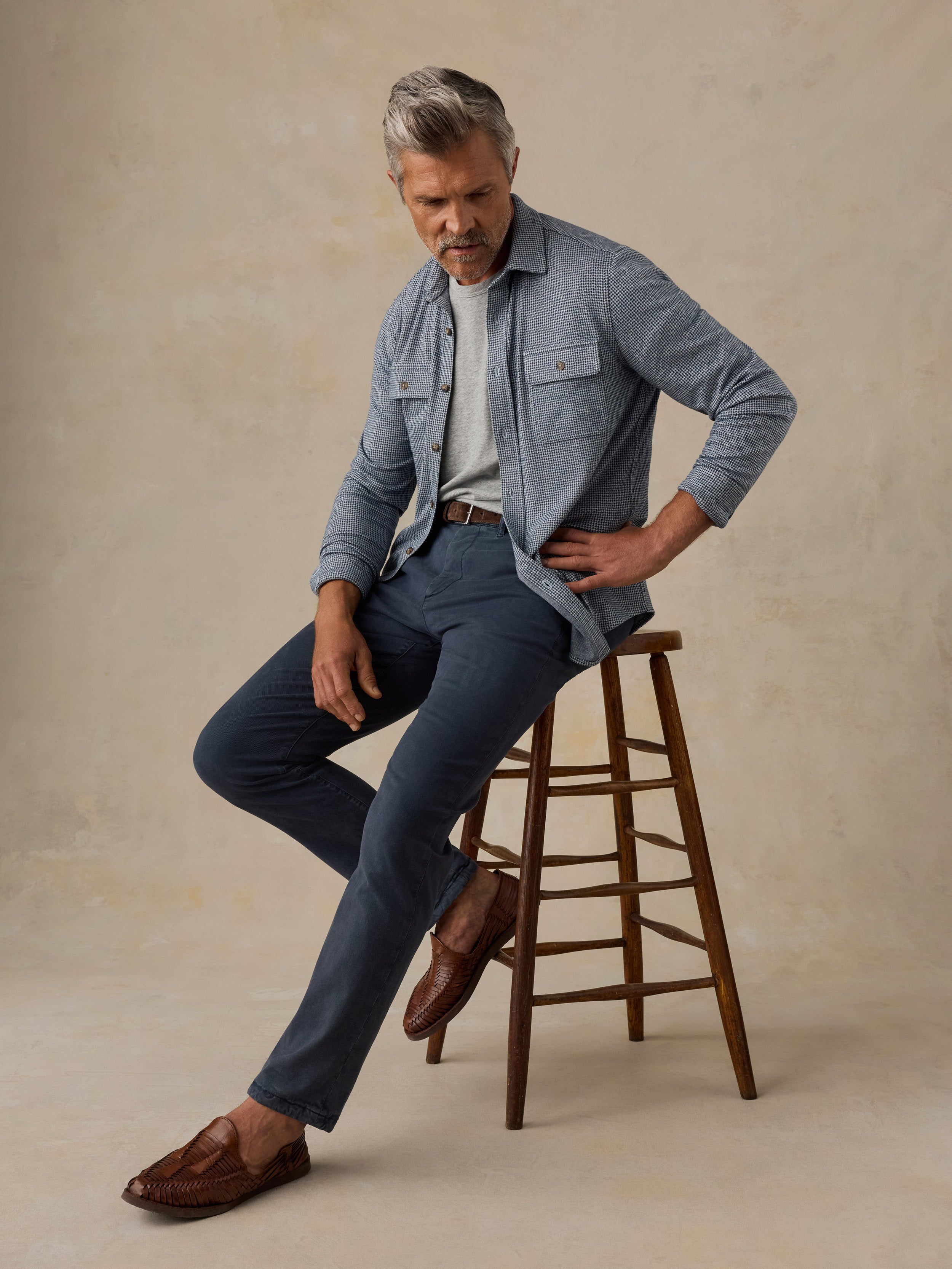 Man sitting on a wooden stool wearing blue pants and a striped sweater on a beige background