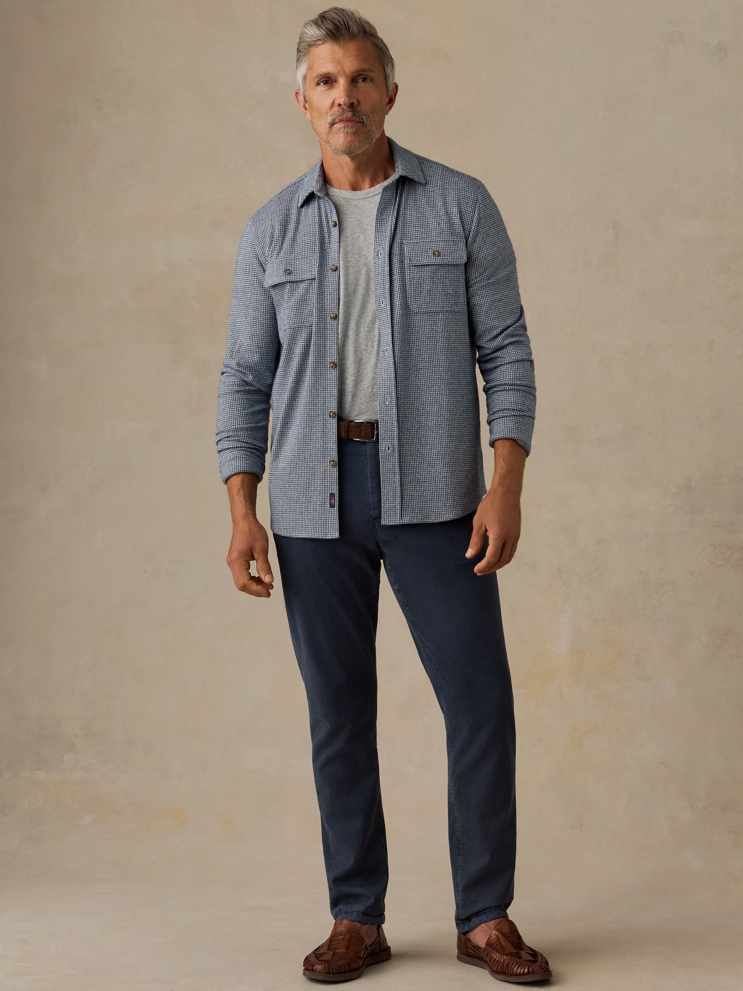 Man wearing a grey sweater shirt and blue pants standing against a beige wall with a wooden chair.