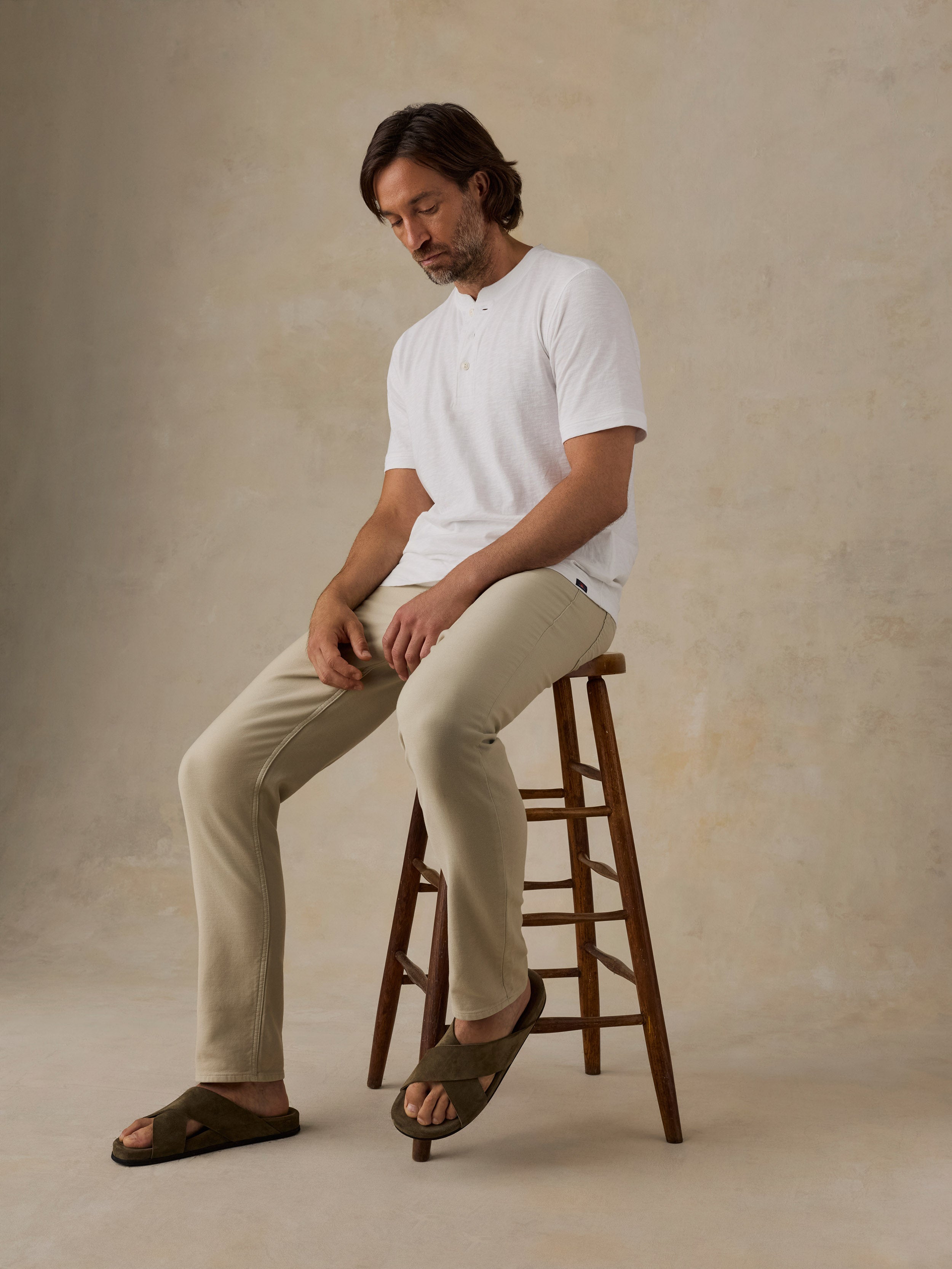A man sits on a wooden stool wearing a white short-sleeve henley shirt, beige trousers, and dark brown slide sandals against a textured beige studio backdrop.