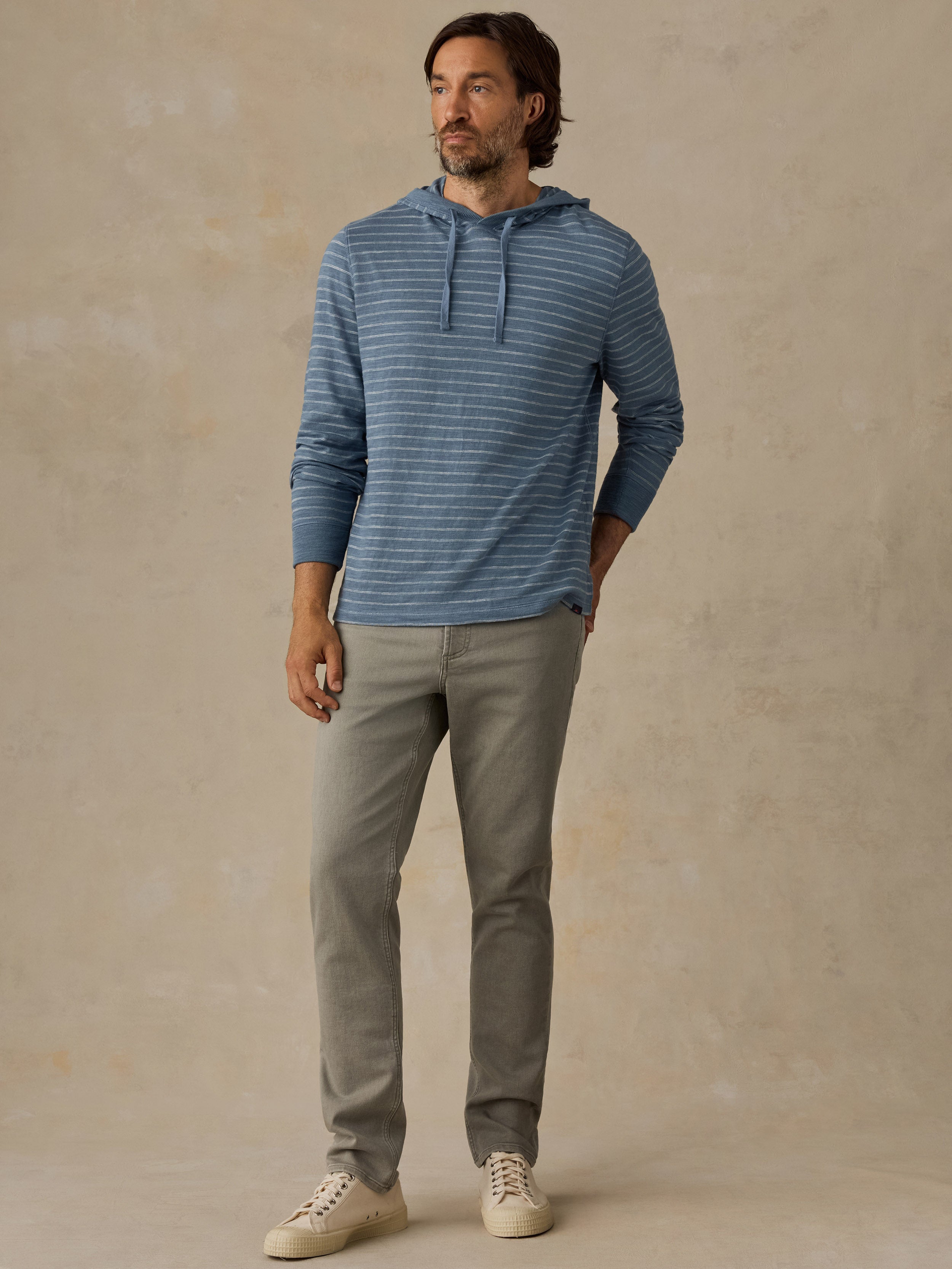 A man wearing a blue striped hoodie, light green slim-fit jeans, and white sneakers, standing against a solid beige studio backdrop.