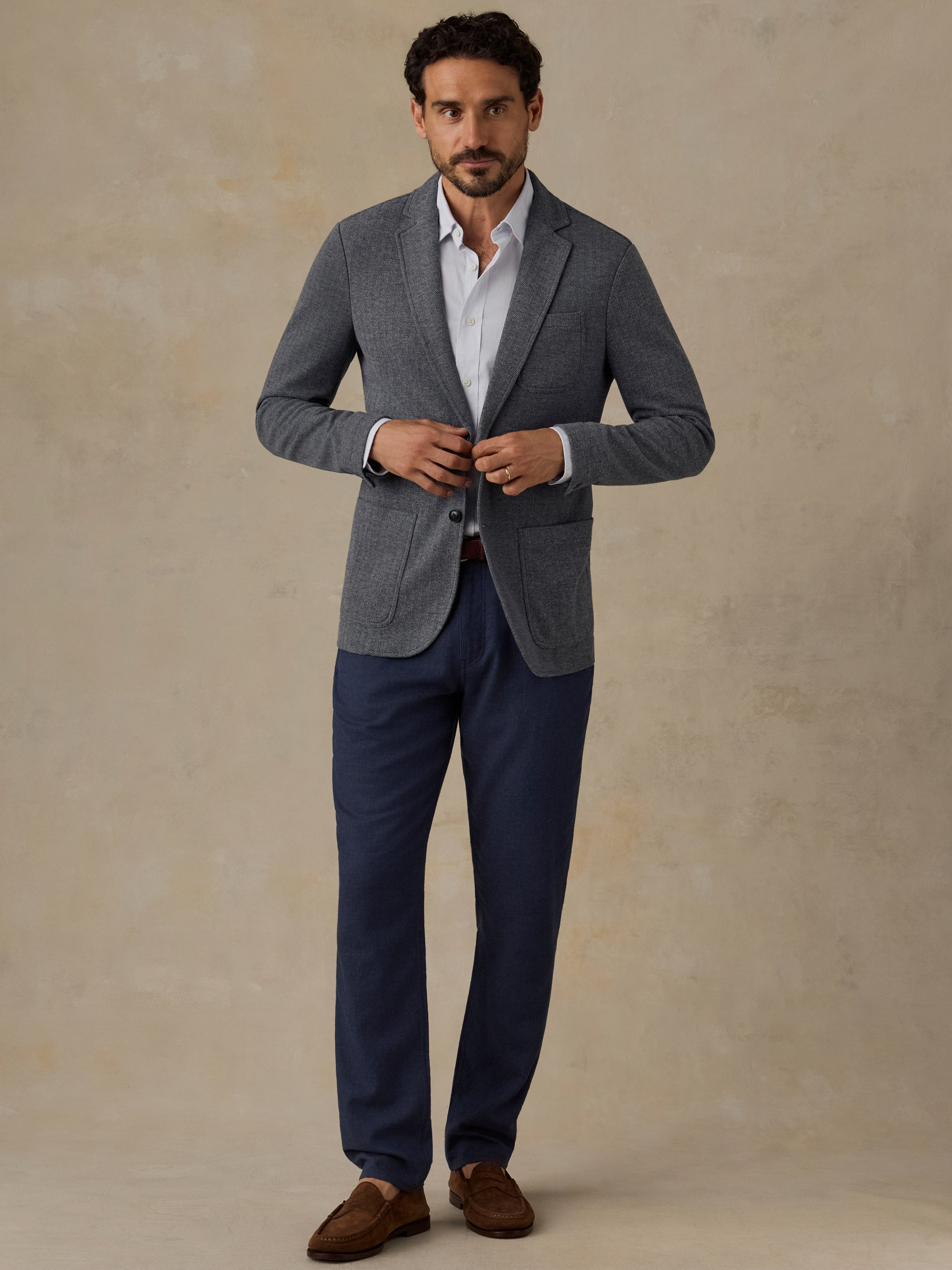 Man wearing a grey blazer and navy blue linen pants with brown loafers against a beige studio background