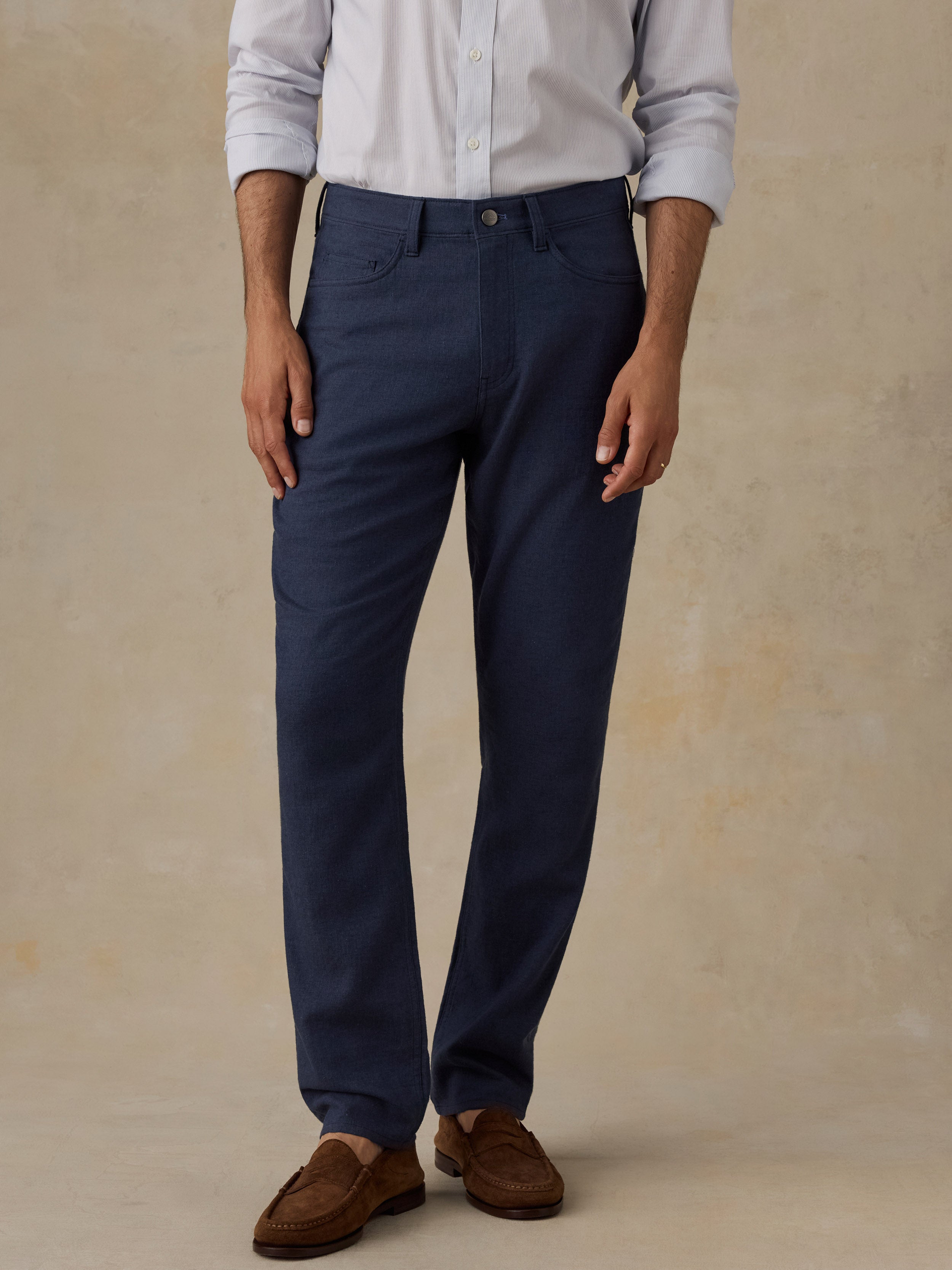 A man wearing navy blue straight-leg dress pants, a light-colored button-down shirt, and brown suede loafers, standing against a textured beige studio backdrop.