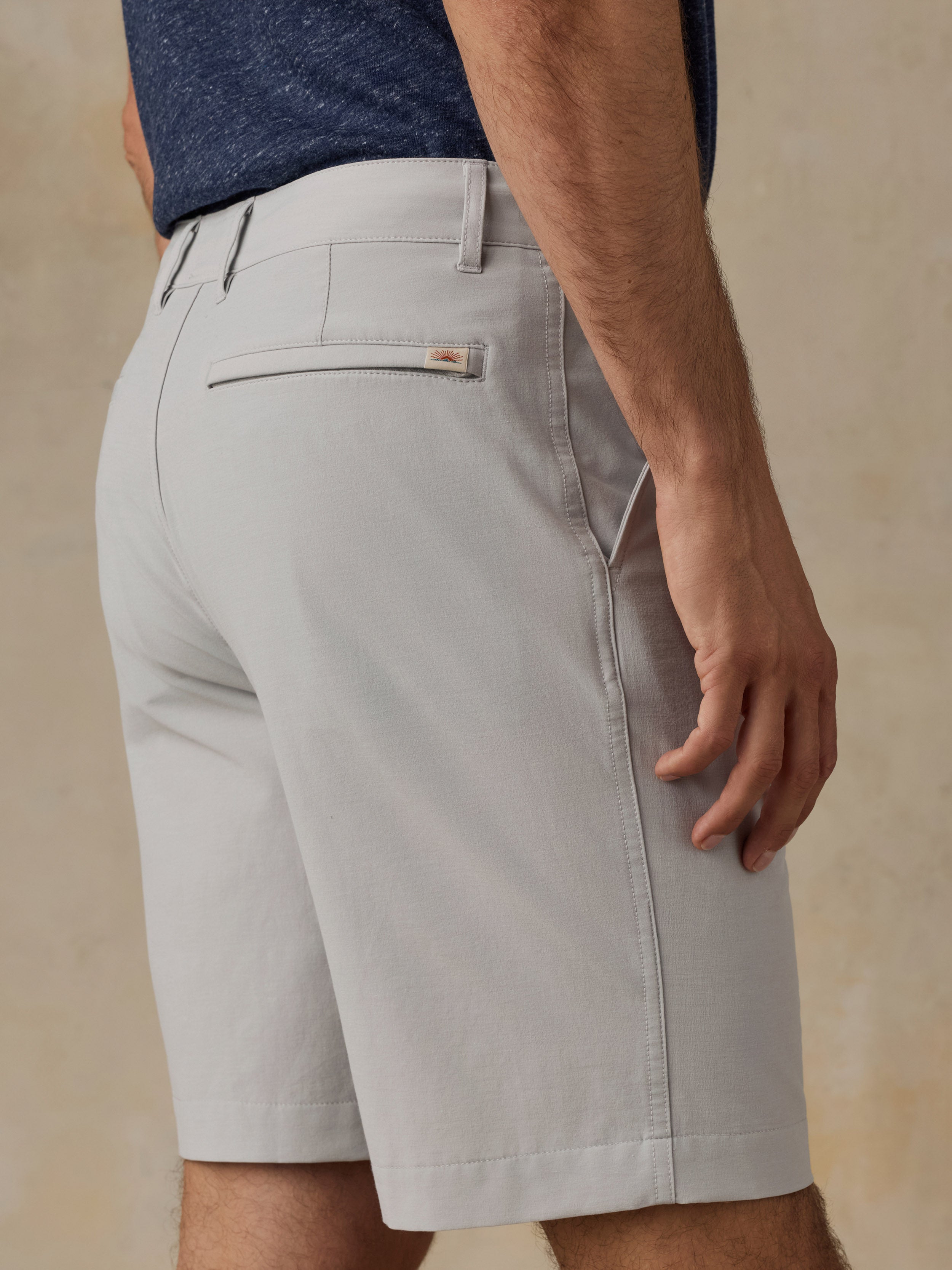 A close-up of a man wearing light gray shorts, shown from the waist down against a warm beige textured background.