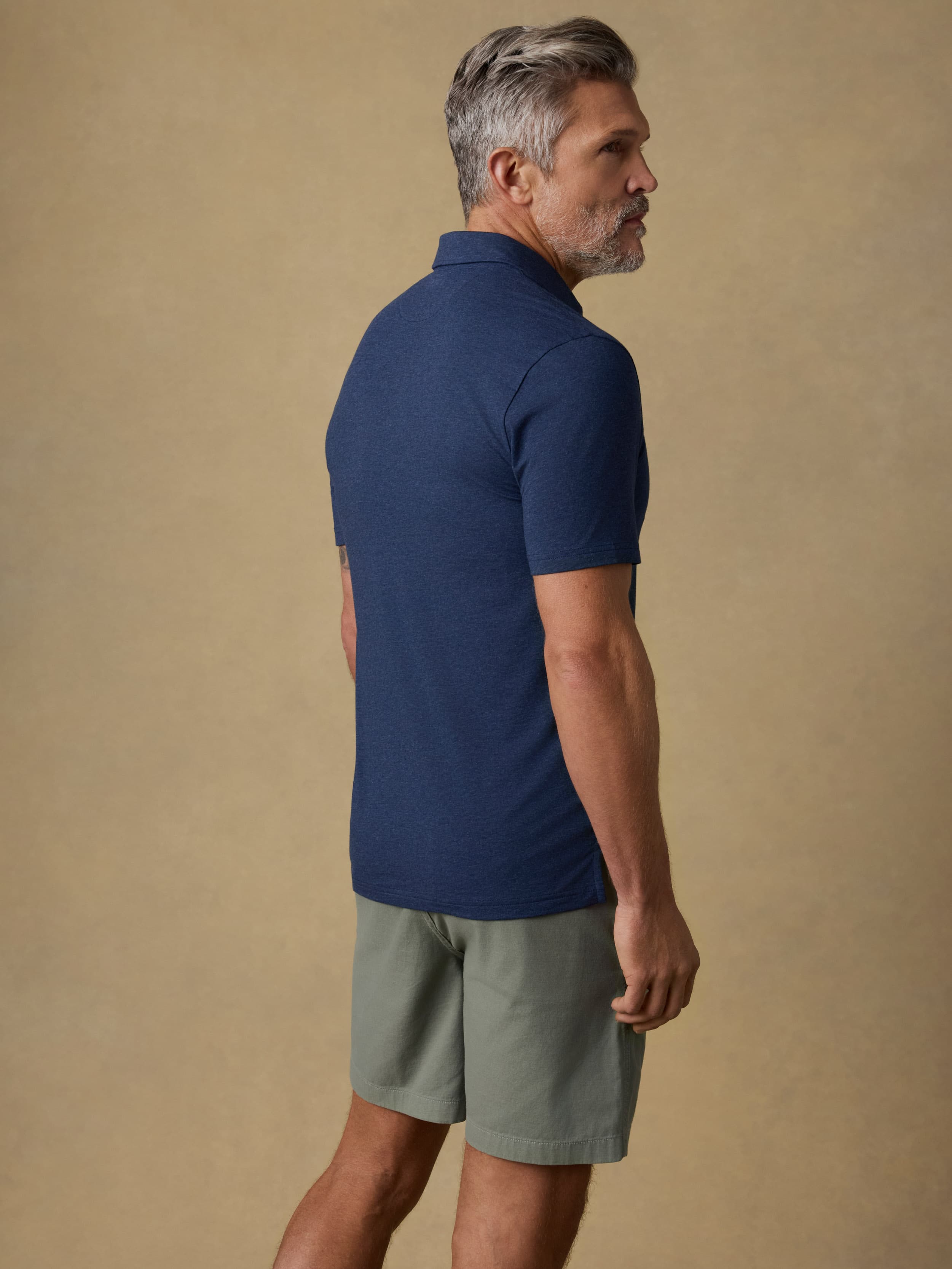 A man wearing a navy blue short-sleeve polo shirt and light olive green shorts, shown from a side-back angle against a solid beige studio background.