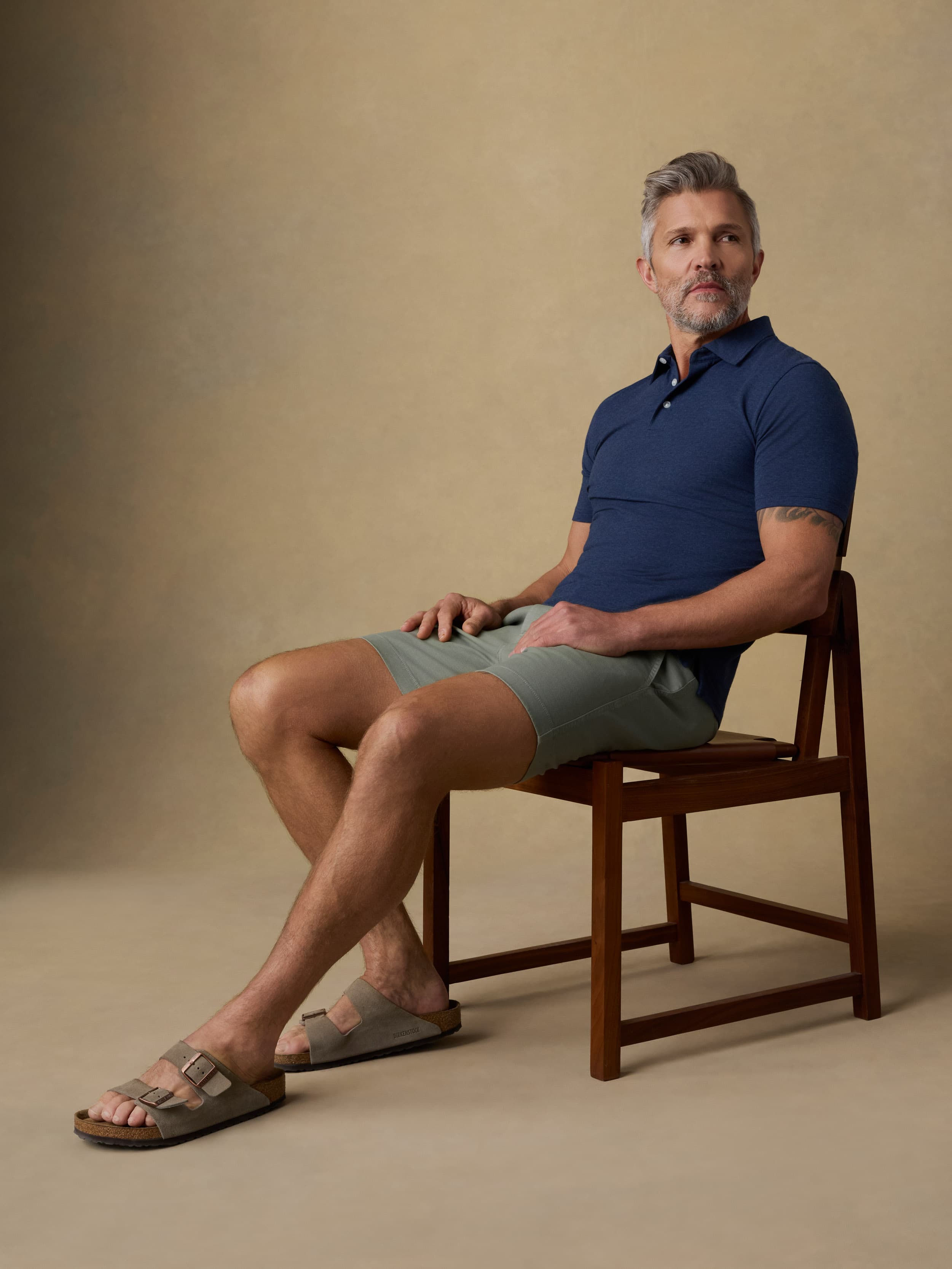 A man sits on a wooden chair wearing a navy blue polo shirt, light olive green shorts, and brown slide sandals against a warm beige studio backdrop.