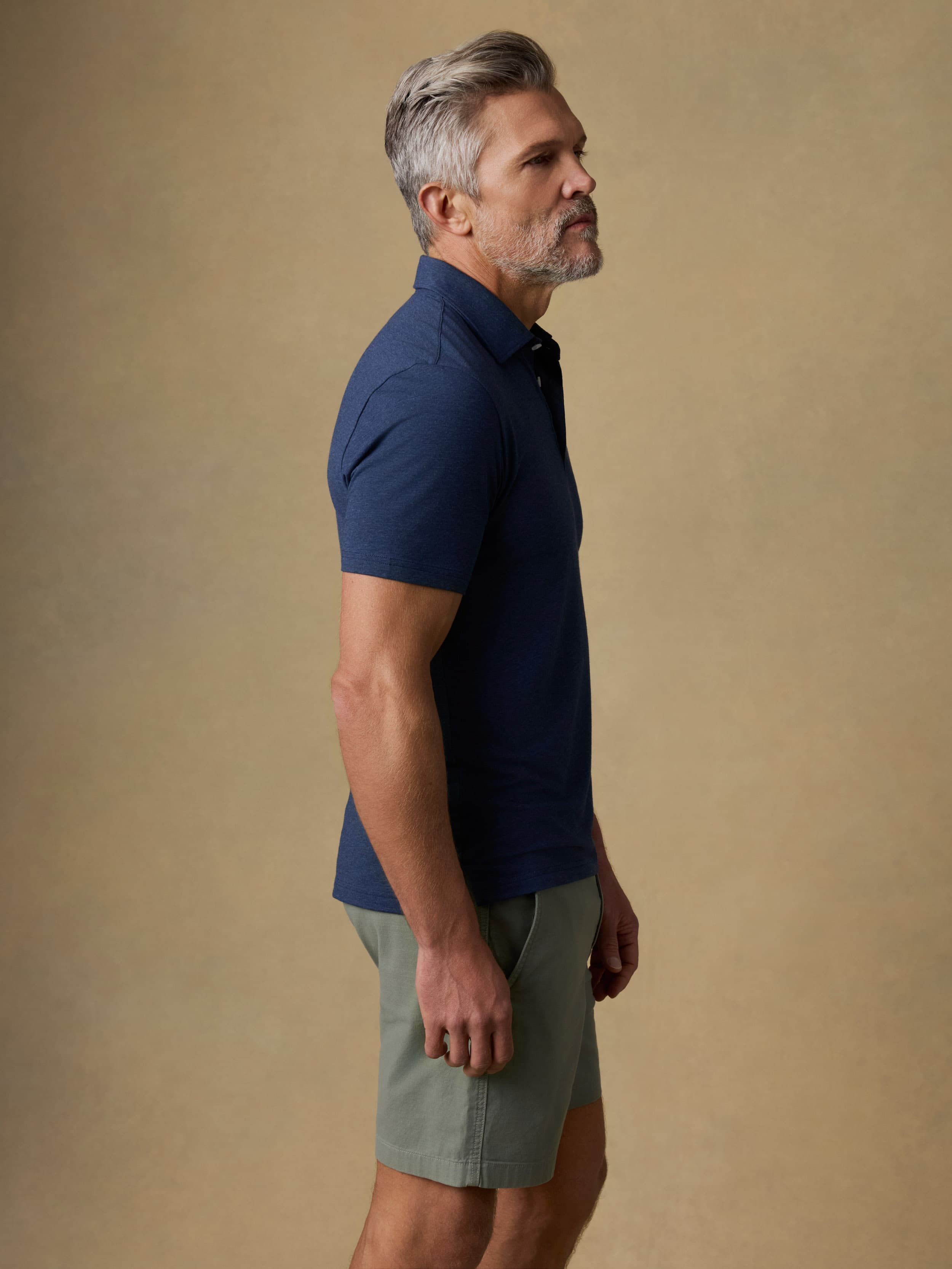 A man in side profile wearing a navy blue short-sleeve polo shirt and light olive green shorts, standing against a solid beige studio background.