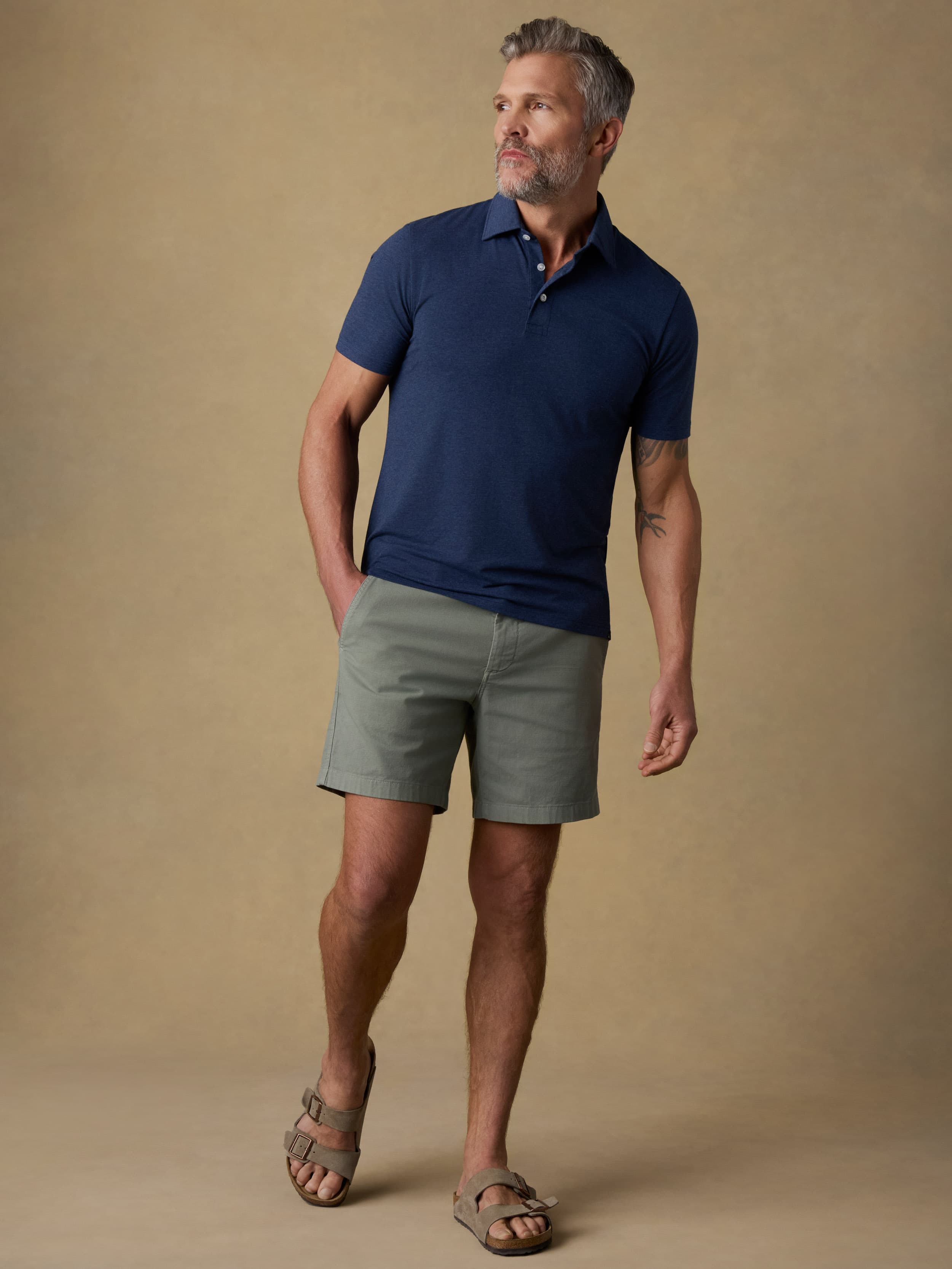 A man wearing a navy blue short-sleeve polo shirt, olive green shorts, and brown sandals, standing against a solid beige studio background.