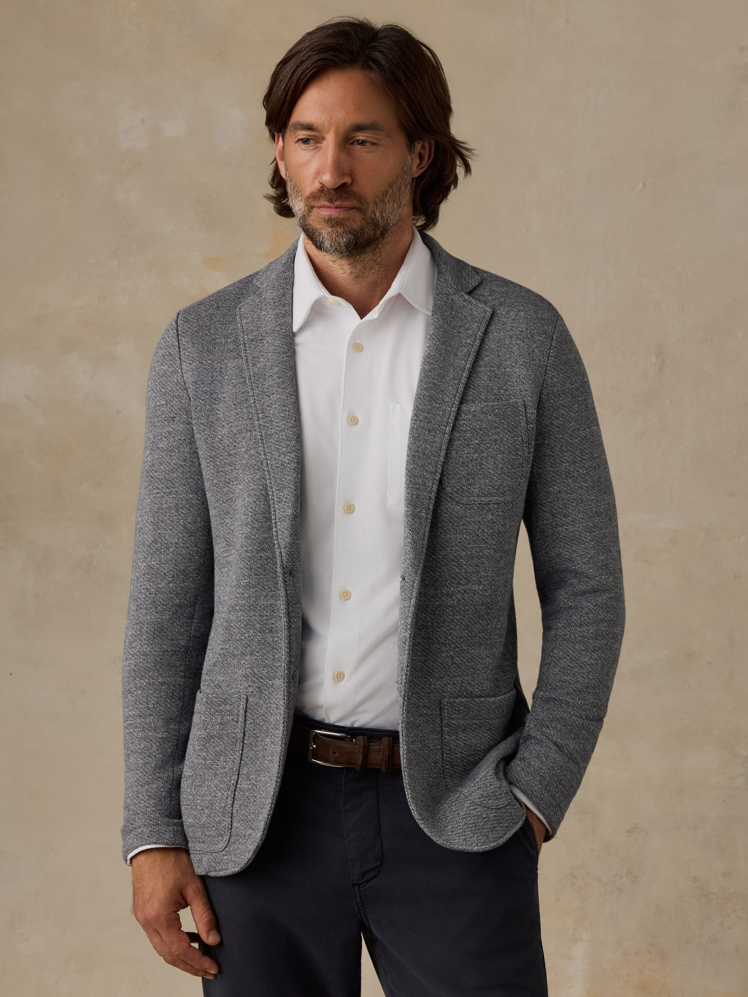 Man wearing a grey blazer over a white button down shirt and navy pants against a beige background