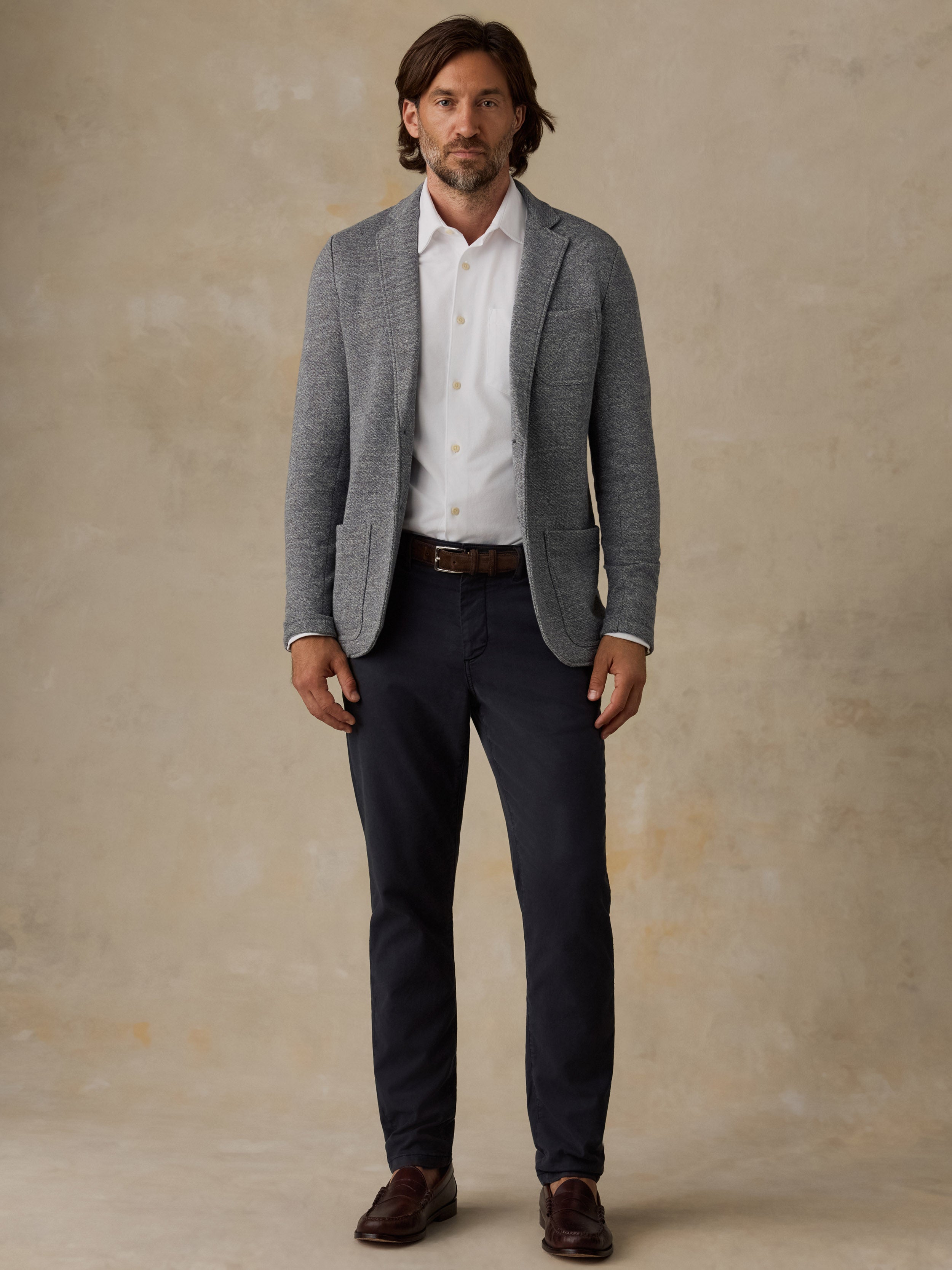 Man wearing a grey blazer over a white button down shirt and navy pants against a beige background
