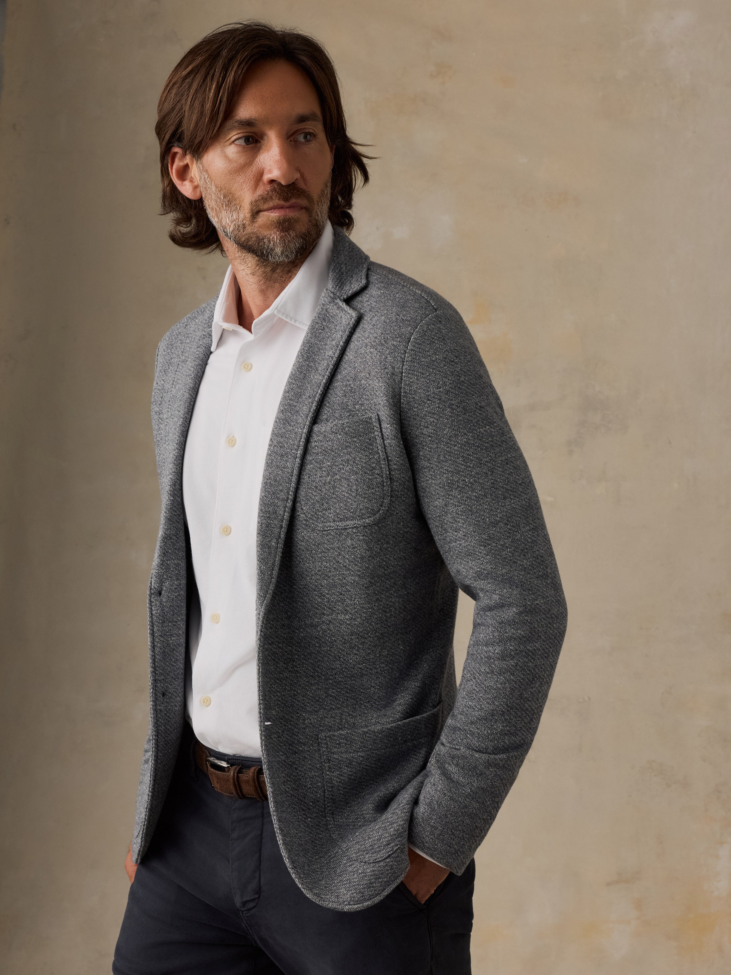 Man wearing a grey blazer over a white button down shirt and navy pants against a beige background