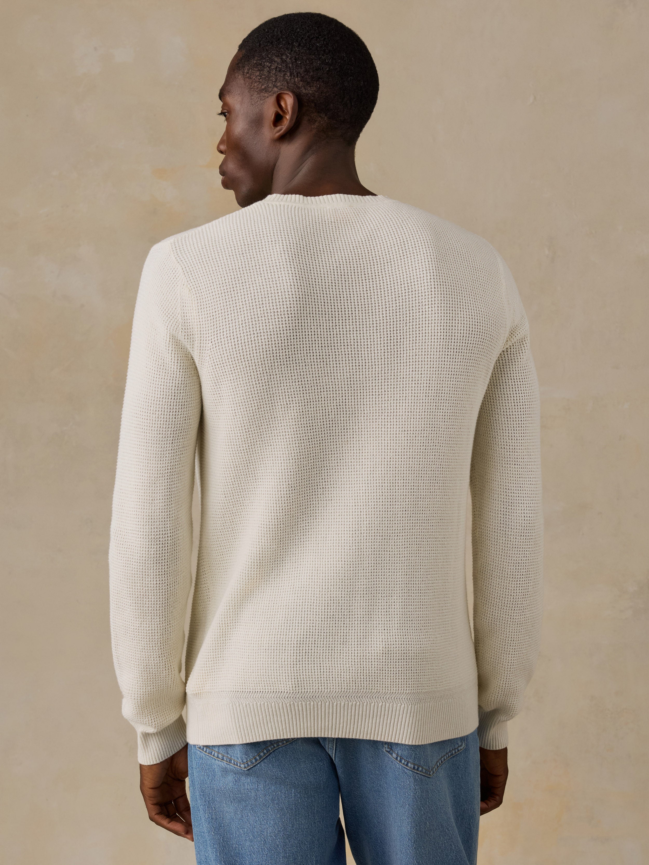 A man wearing a cream-colored ribbed knit sweater and light blue patterned shorts, shown from a side-back angle against a solid beige background.