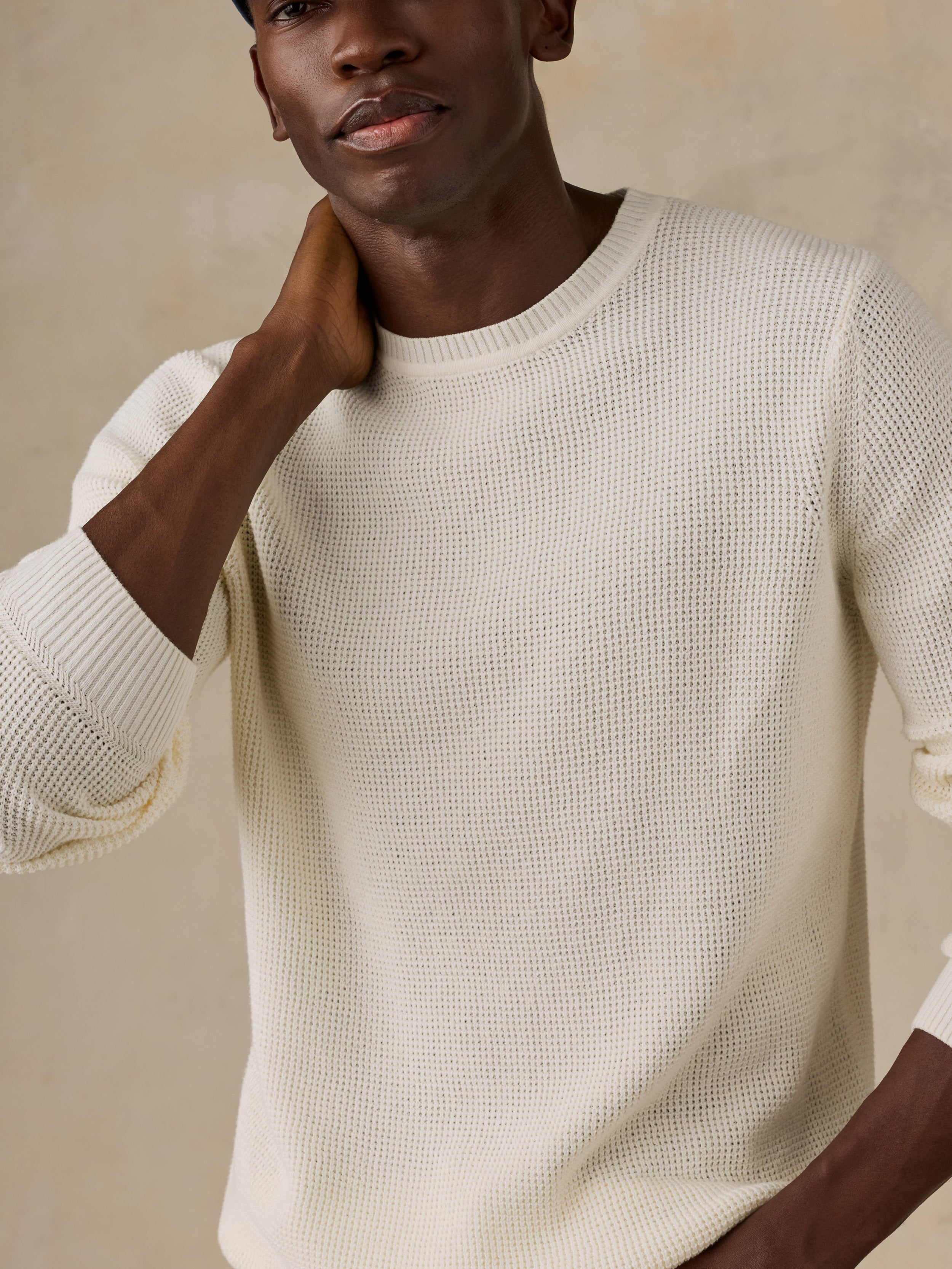 A close-up of a man wearing a textured cream waffle-knit crewneck sweater against a warm beige background.