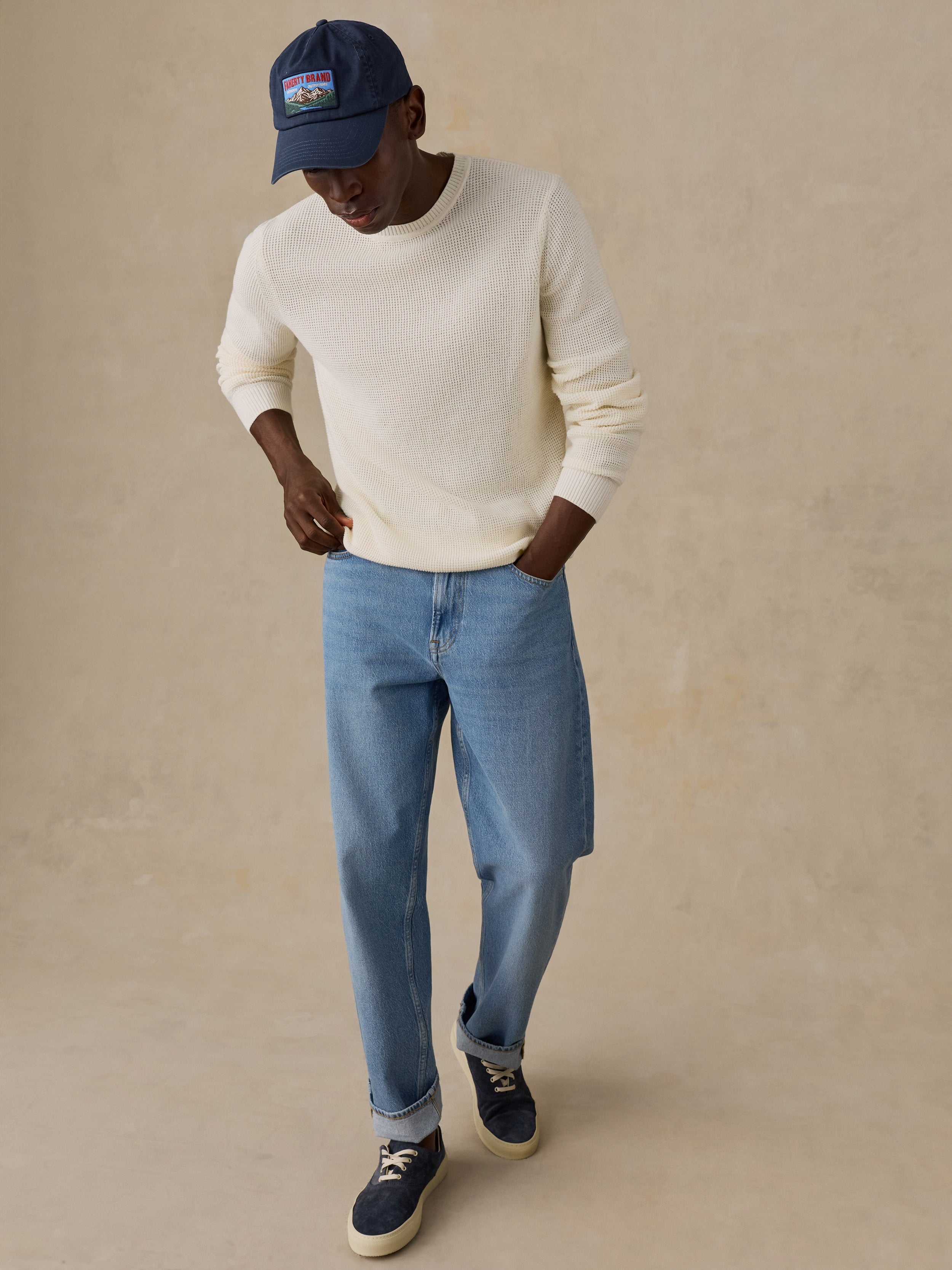 A man wearing a cream-colored waffle-knit crewneck sweater, standing against a warm beige background.