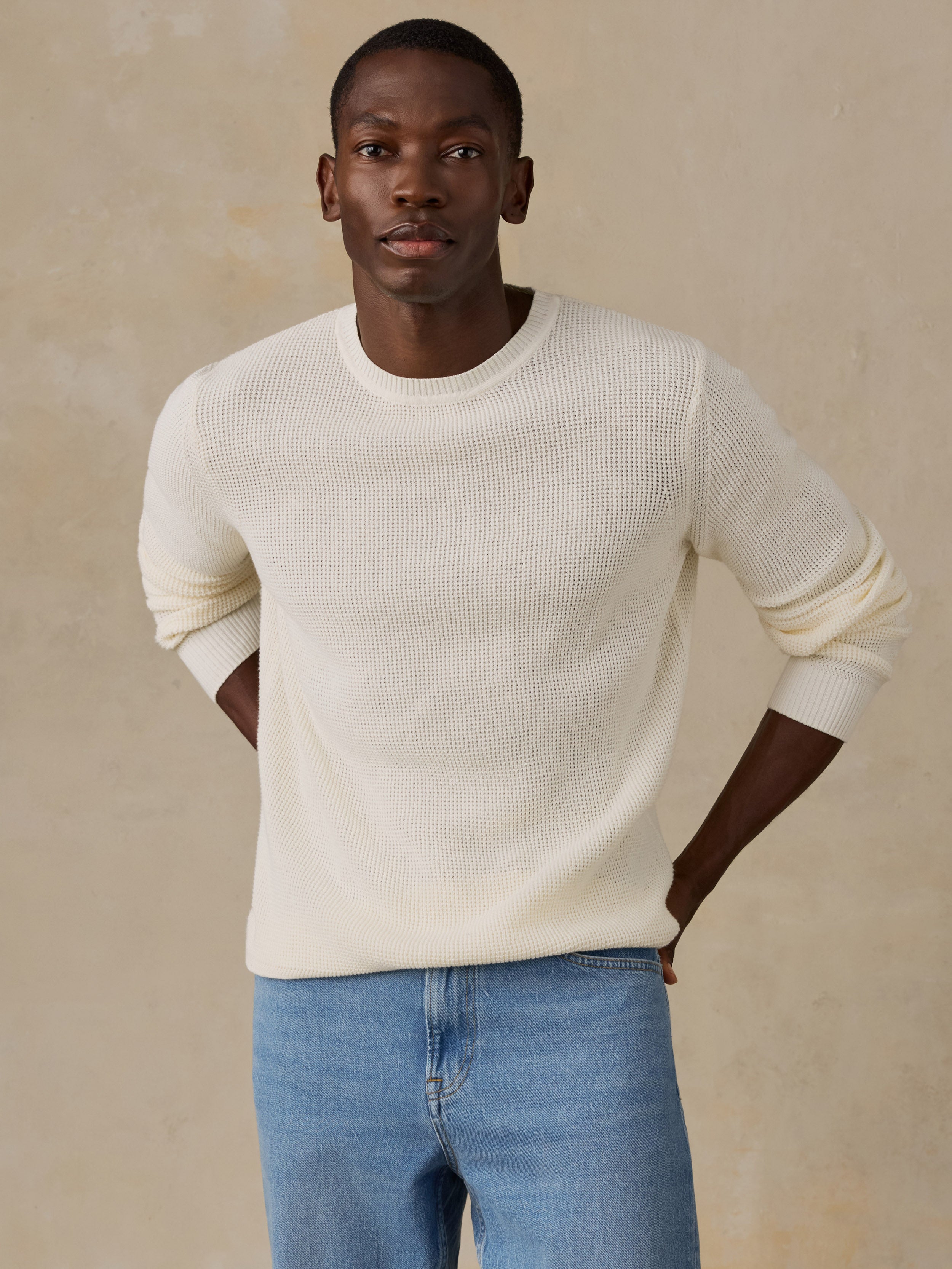 A man wearing a cream ribbed knit sweater and light blue patterned shorts, standing against a warm beige studio background.