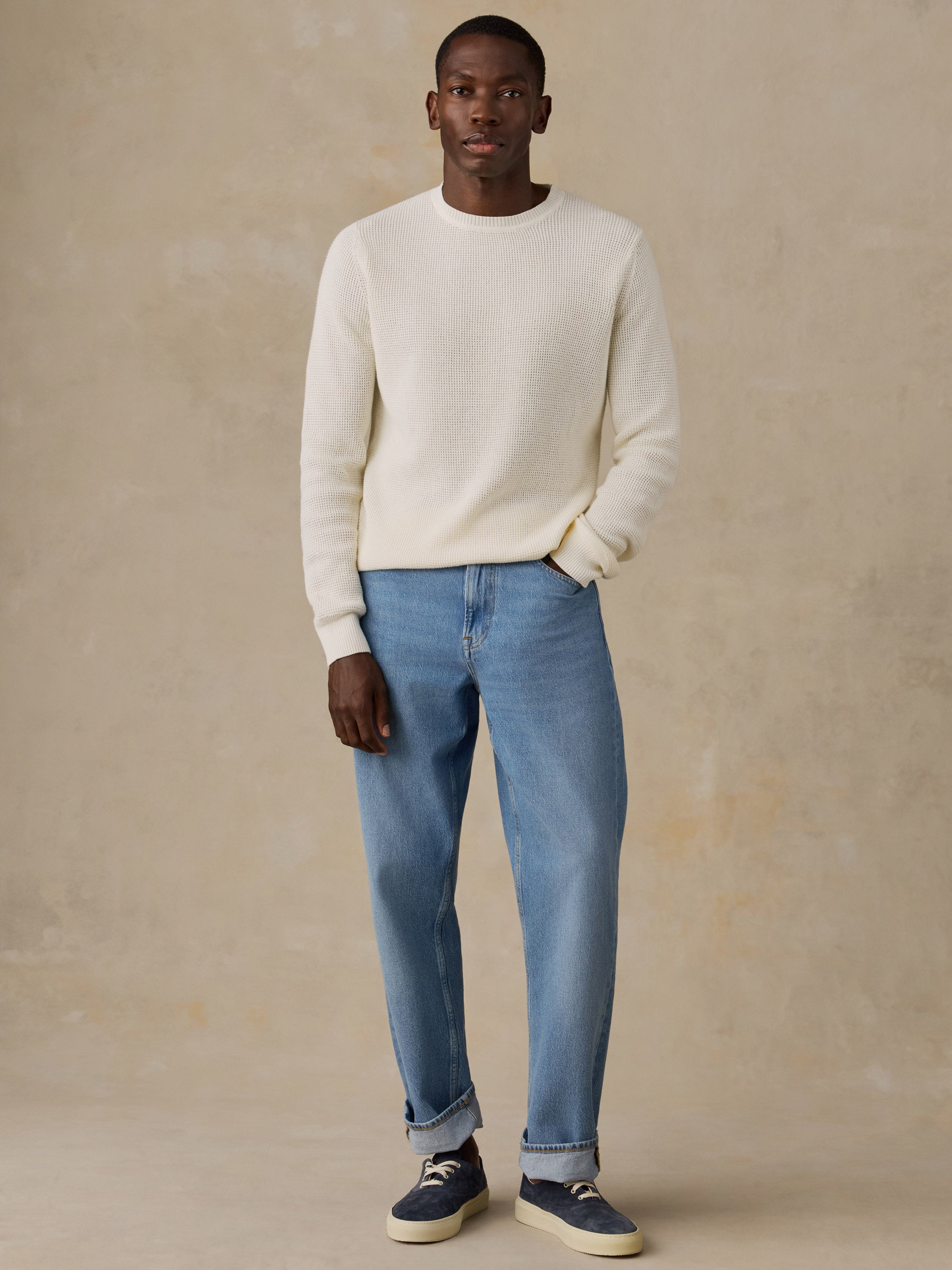 A man wearing a cream knit crewneck sweater, light blue patterned shorts, and white sneakers, standing against a solid beige studio background.