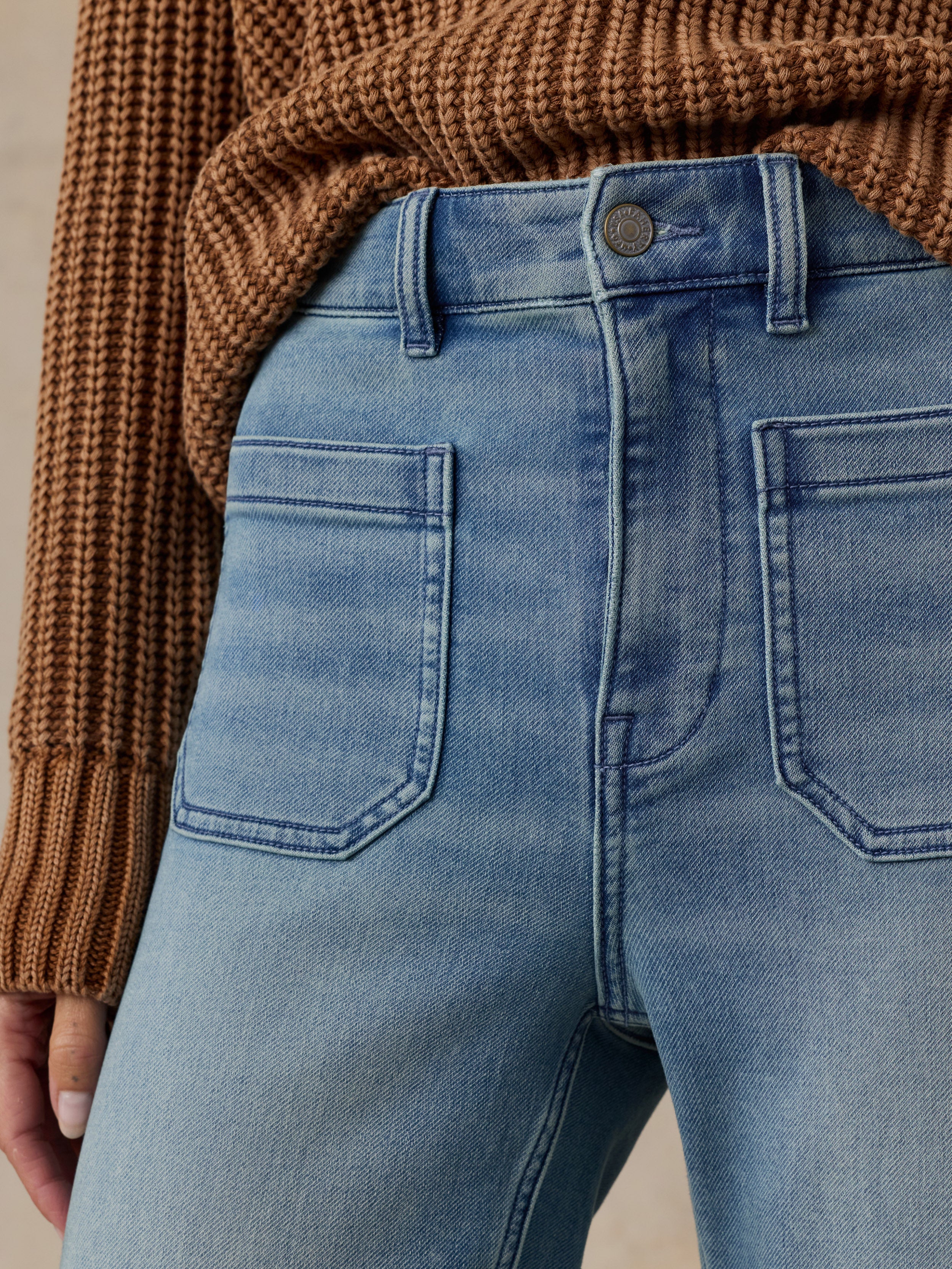 Close-up of the waistband and pockets of light blue denim jeans, styled with a brown ribbed knit sweater.