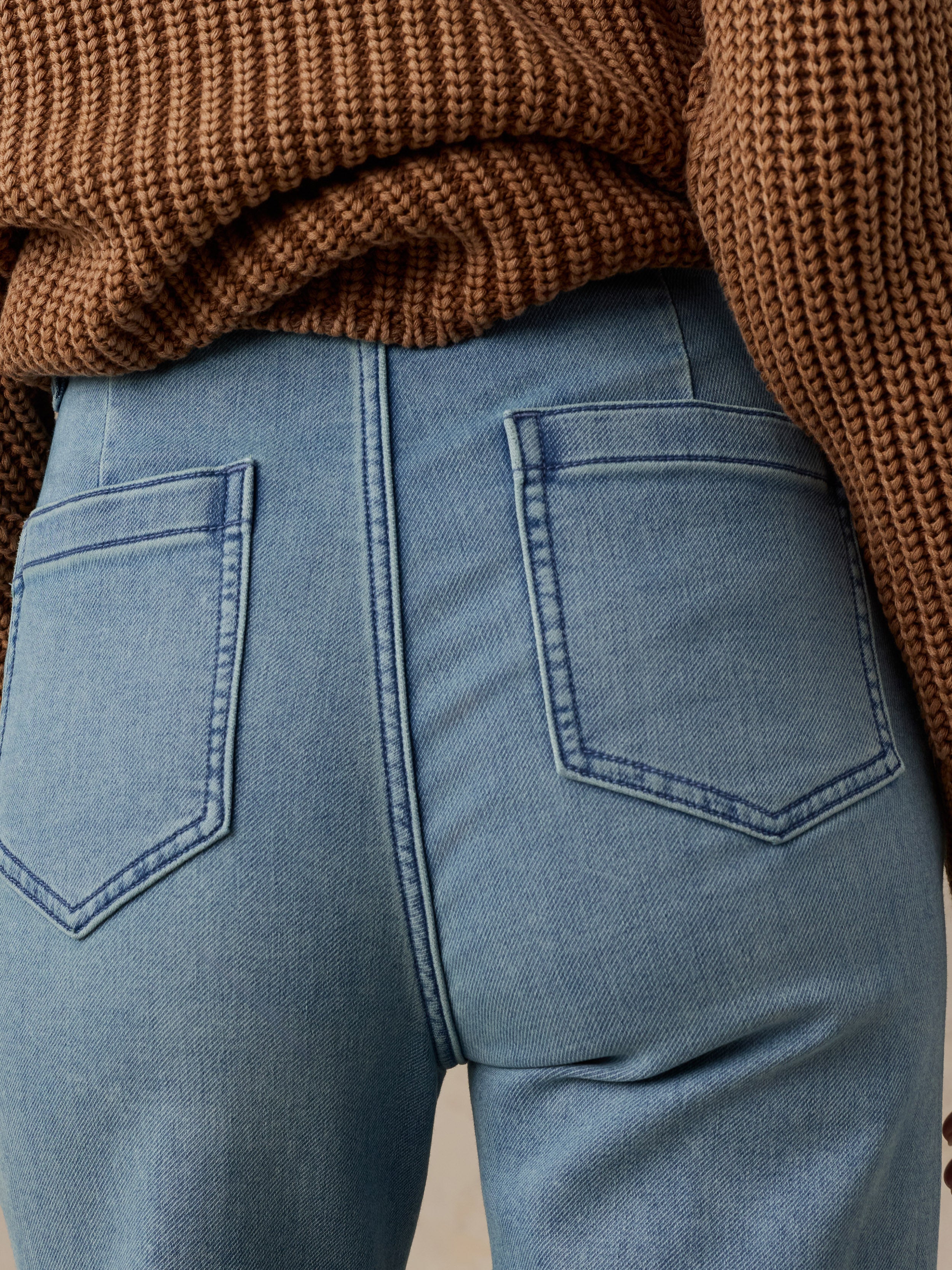 Rear view of a woman wearing light blue denim jeans with a ribbed light pink top, set against a solid beige background.