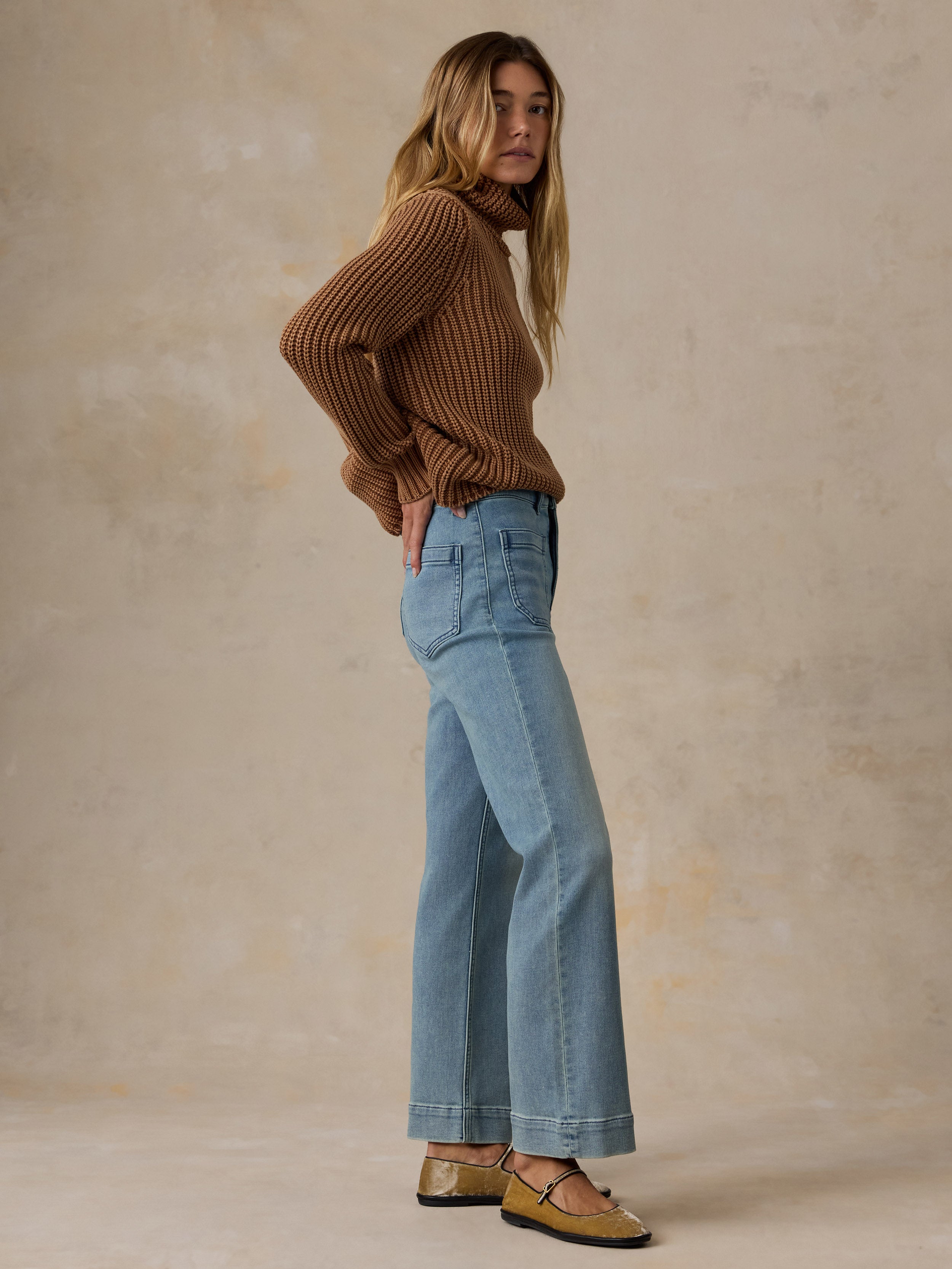 A woman modeling a pair of high-waisted, wide-leg cropped blue jeans with a white tank top and brown flat shoes, standing against a textured beige studio backdrop.