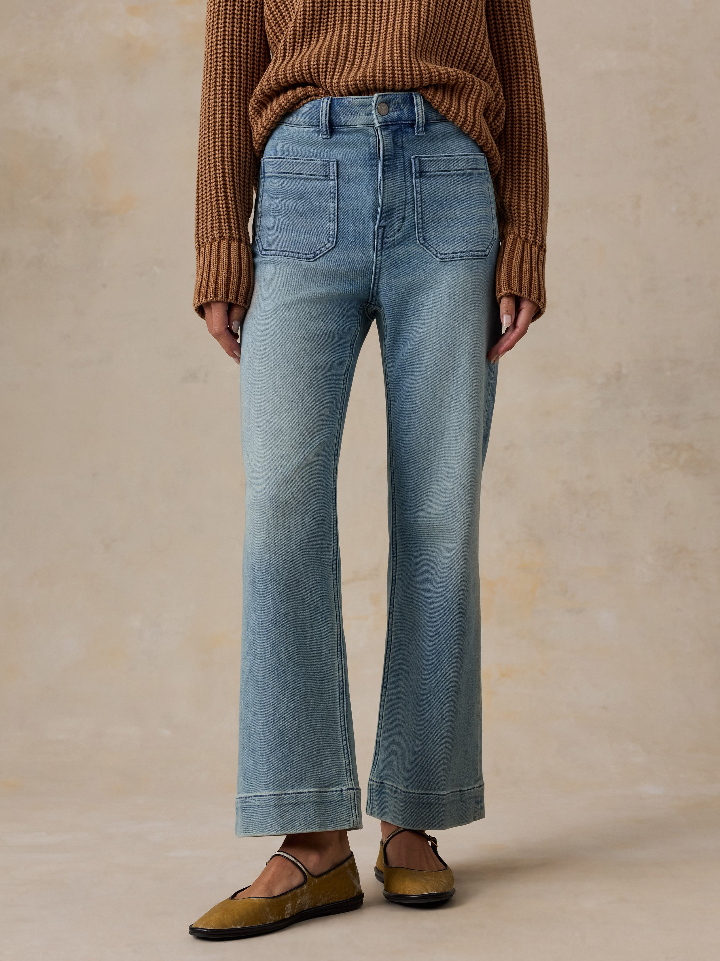 A person wearing light-wash blue flare jeans with a brown ribbed sweater and brown shoes, standing against a warm beige textured studio background.
