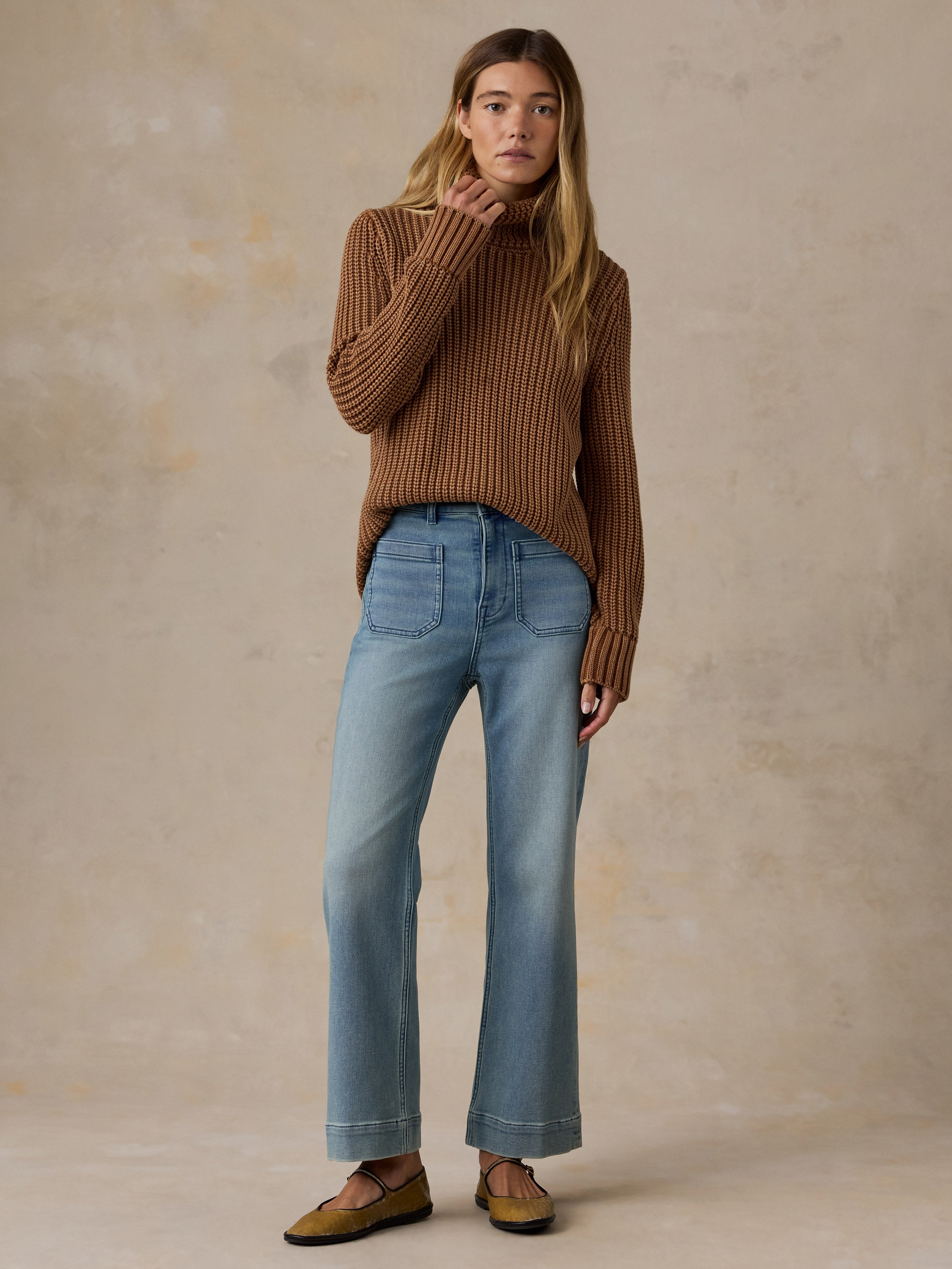 A woman stands against a warm beige studio backdrop wearing a white sleeveless top, light blue cropped flare jeans, and brown mule shoes.
