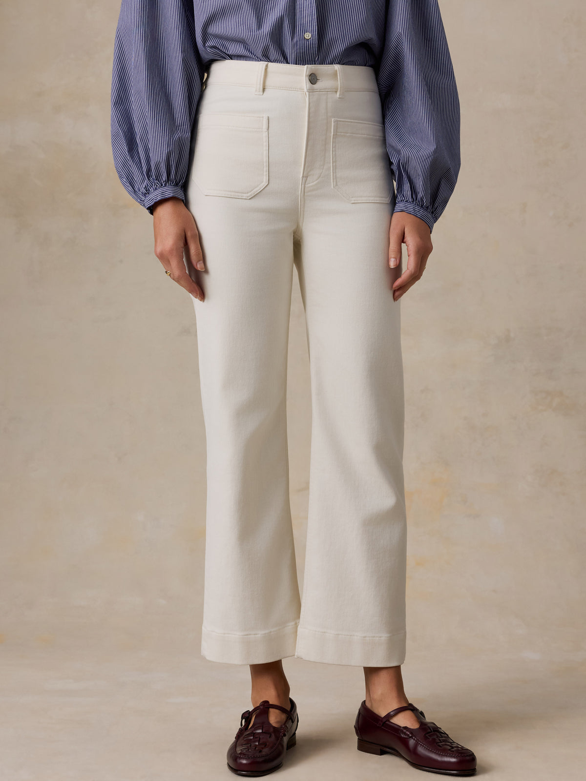 A model is shown from the waist down wearing a pair of off-white cropped wide-leg jeans, a blue striped long-sleeve blouse, and dark burgundy Mary Jane shoes, set against a textured beige studio background., image 2 of 6