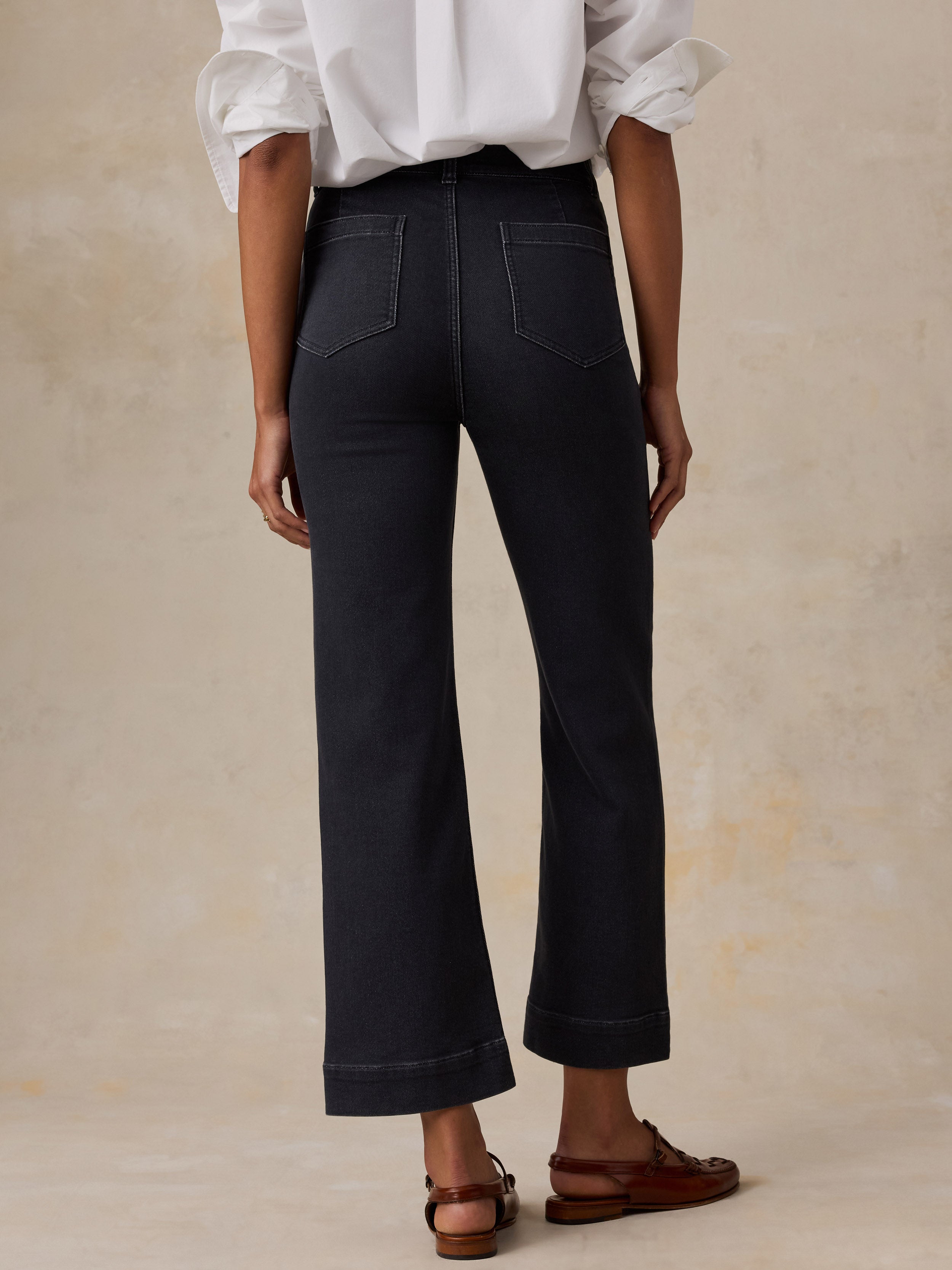 A model shown from the back wearing high-waisted, wide-leg black jeans, a white button-down shirt and brown loafers against a solid beige studio background.