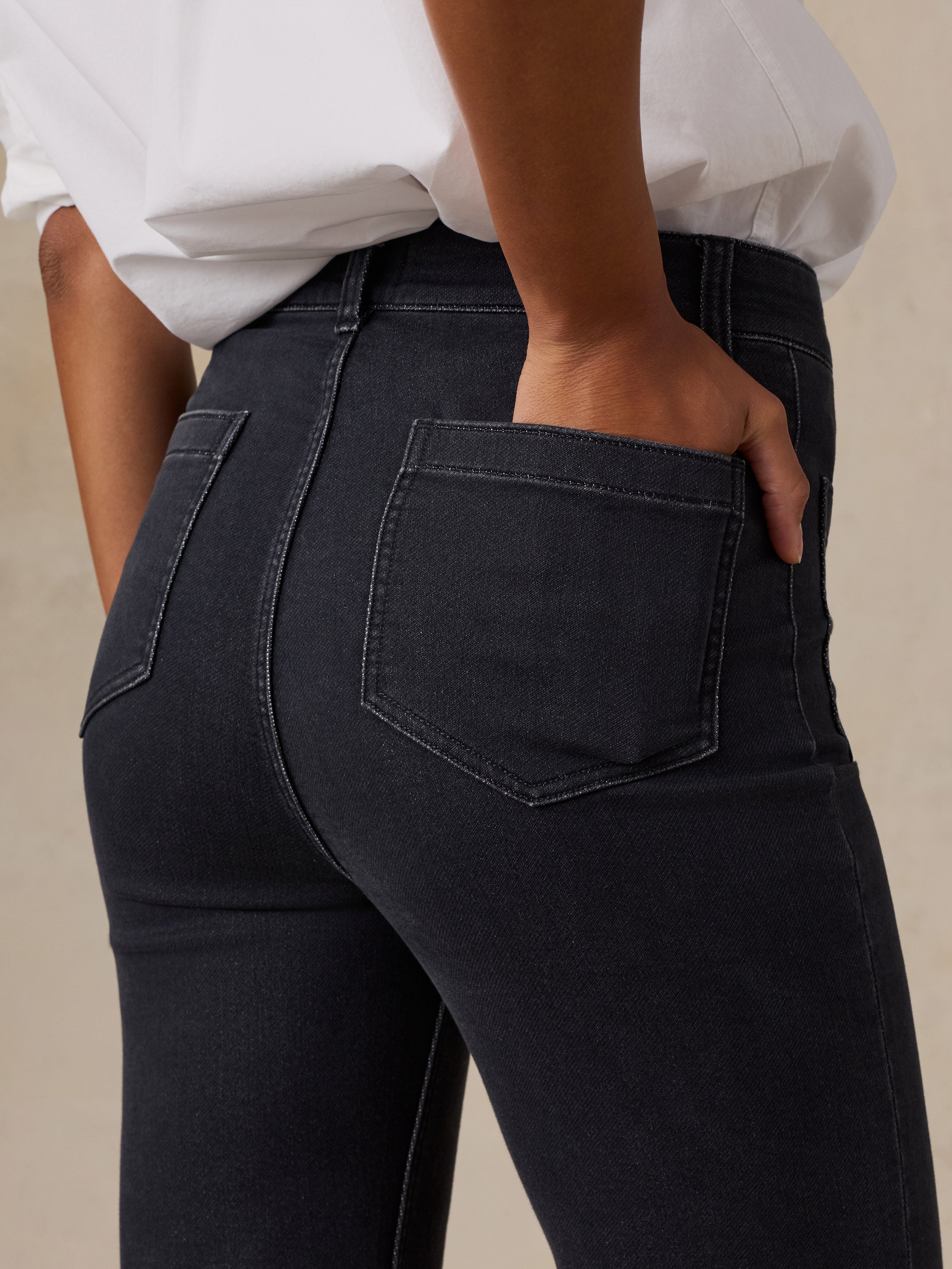 Close-up back view of a woman wearing dark charcoal gray slim-fit jeans with a white button-down shirt tucked in, showing the back pocket detail.